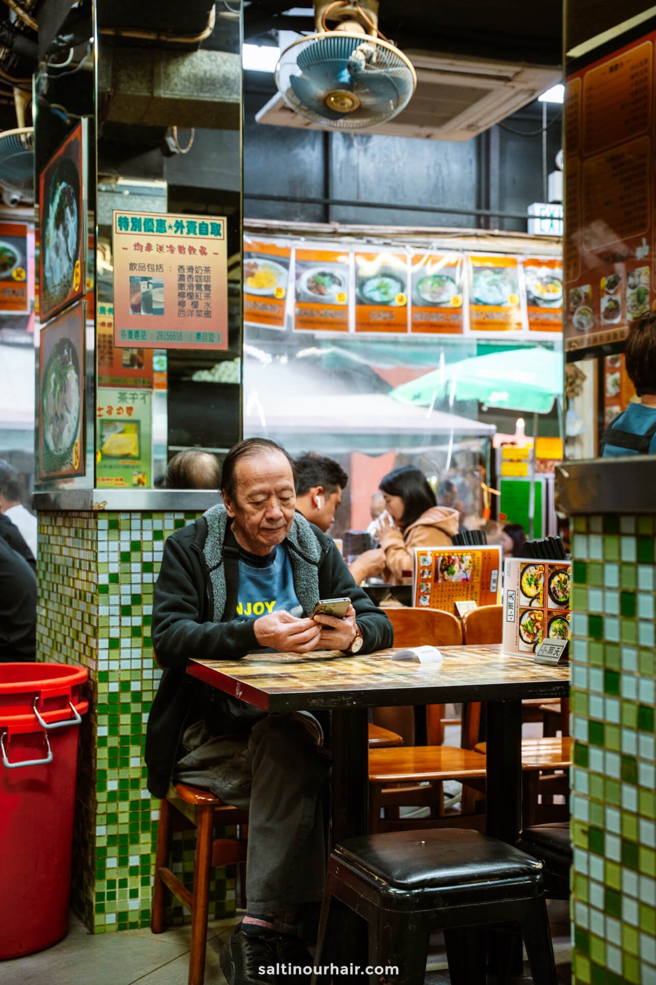An older man sits alone at a tiled table in a casual restaurant, looking at his phone. Menus and food images are displayed on the walls around him.