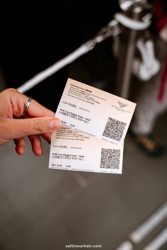 A hand holding two printed tickets with QR codes, against a blurred background of a ticket queue and barrier