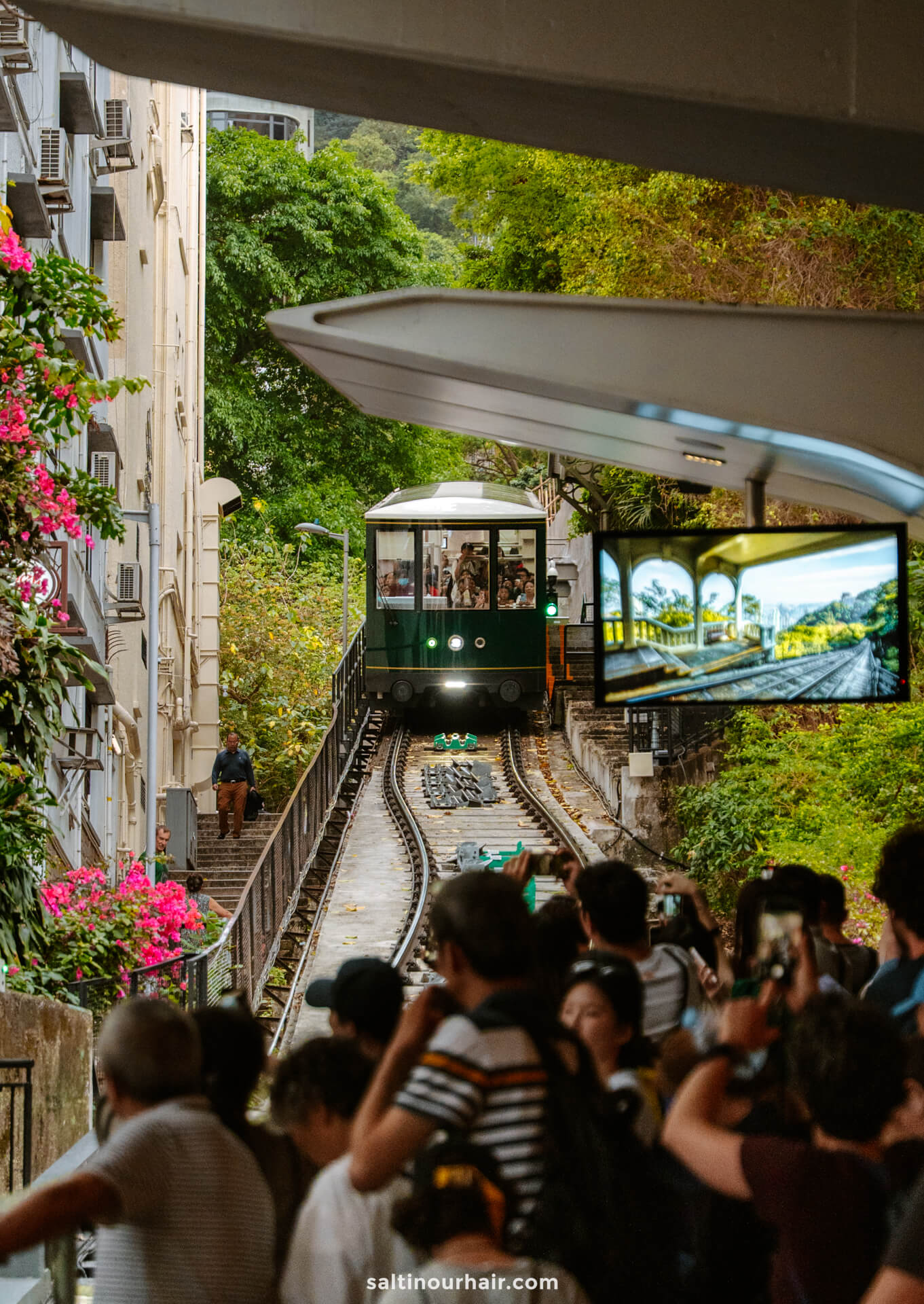 A green tram climbs a steep track between buildings as a crowd watches and takes photos, making it a must-see highlight on any Hong Kong itinerary.