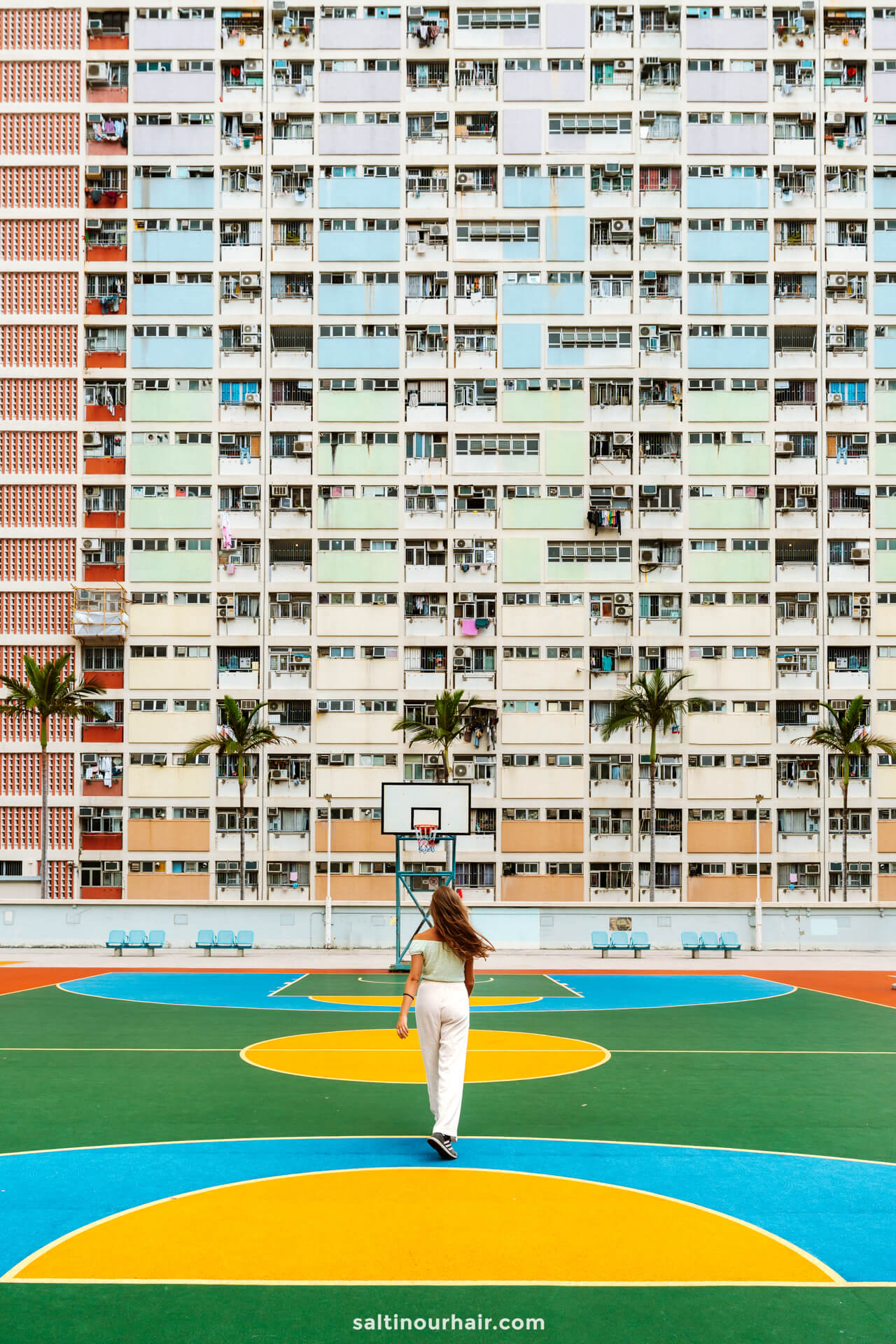 A person walks on a colorful basketball court in front of a tall, multi-colored apartment building with balconies and palm trees&mdash; called Choi Hung estate in Hong Kong