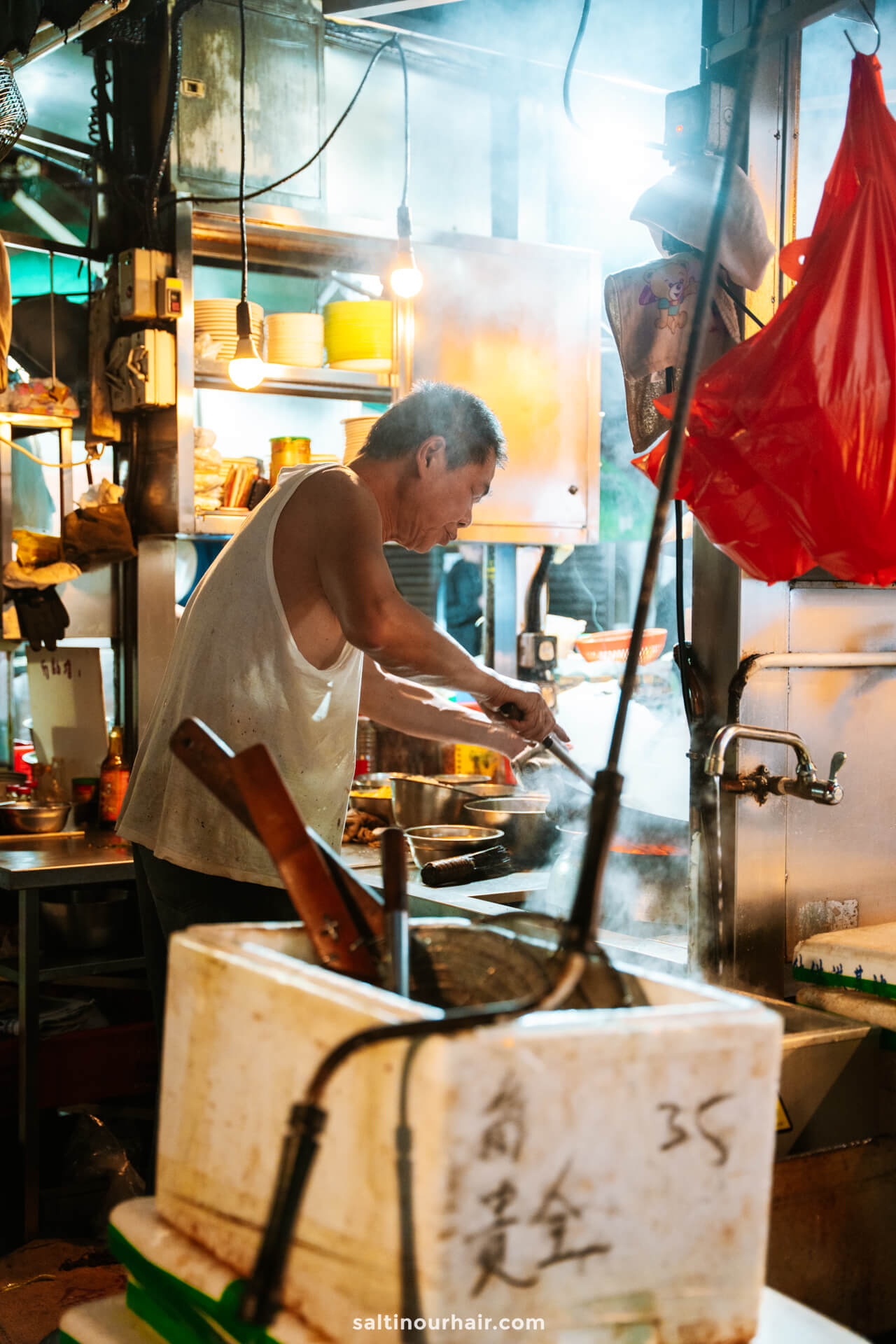 A man in a white tank top cooks food at a busy street food stall in Central Hong Kong, surrounded by steam, utensils, supplies, and hanging light bulbs