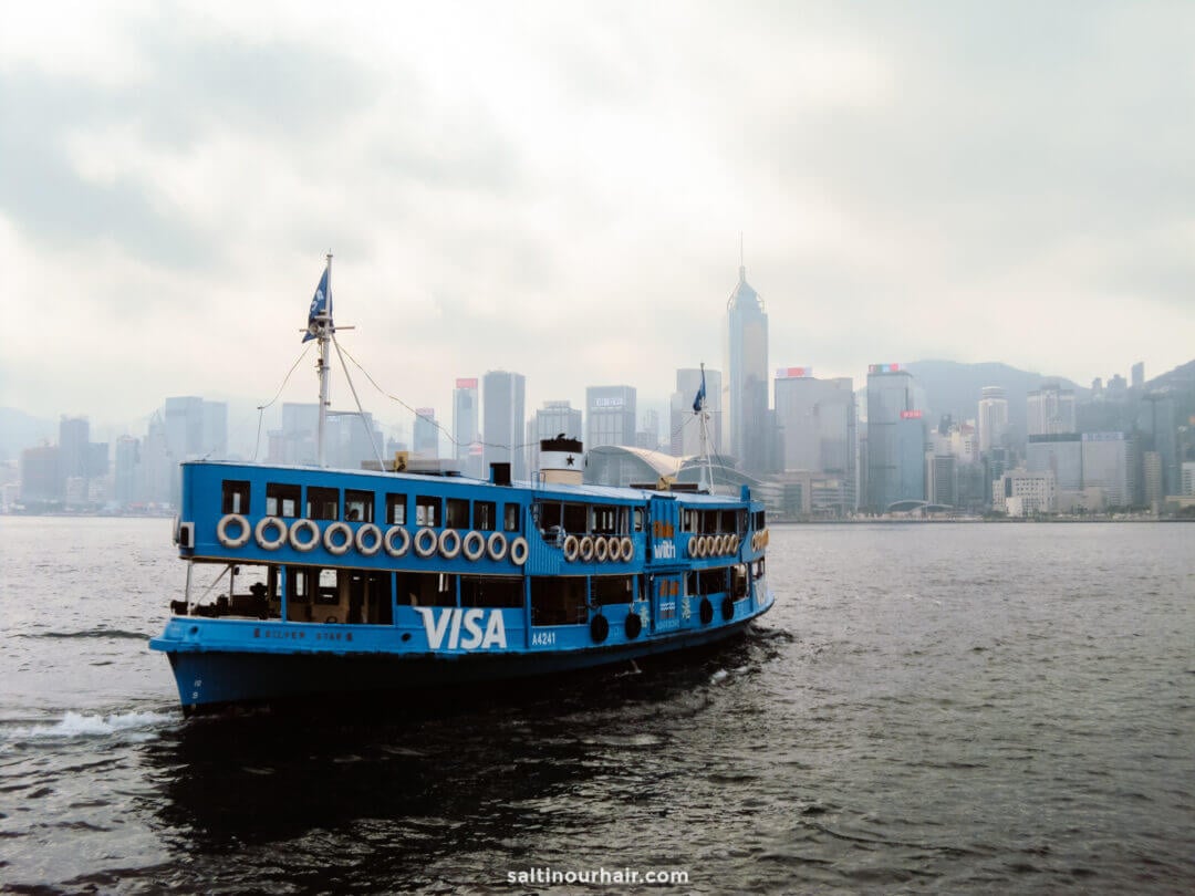 A blue ferry sails on water, the city skyline and tall buildings visible through mist&mdash;a classic scene to include in your Hong Kong itinerary.