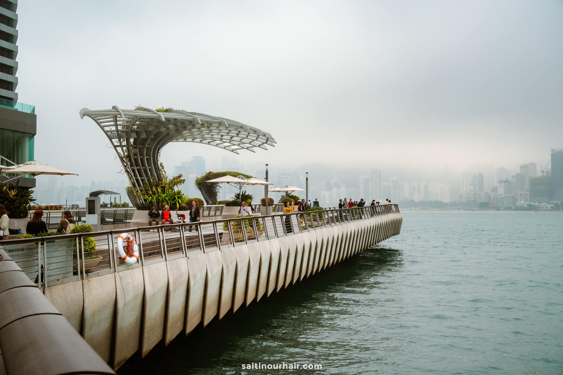 Modern waterfront promenade with people walking and relaxing, perfect for any Hong Kong itinerary, offering views of a misty city skyline across the water.