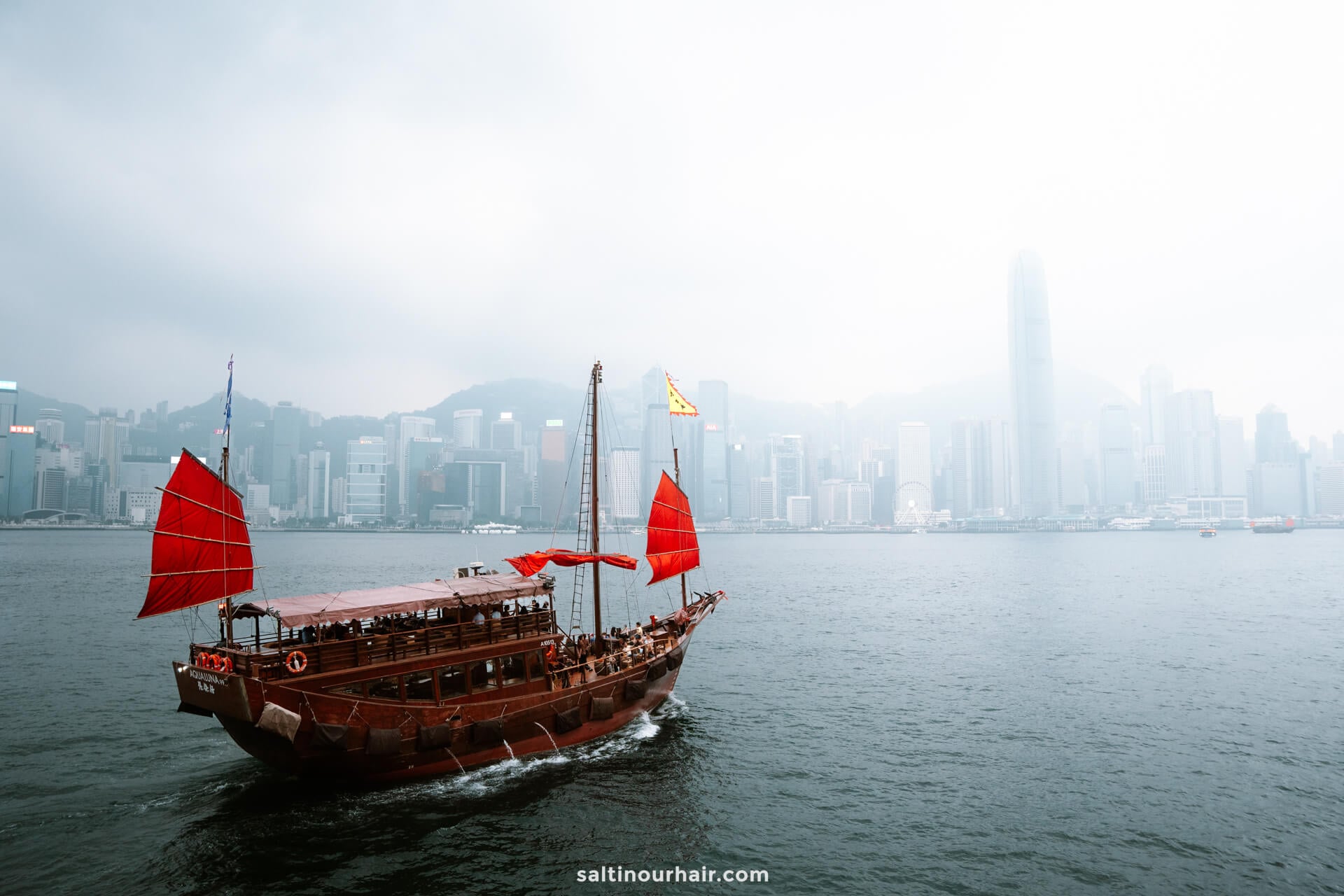 A traditional red-sailed junk boat glides across the water with Hong Kong&rsquo;s modern city skyline in the background