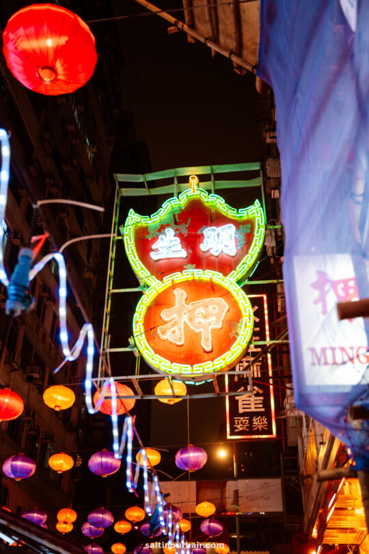 Colorful neon signs and hanging lanterns illuminate a bustling street at night, with buildings and shop signs visible in a market in Hong Kong