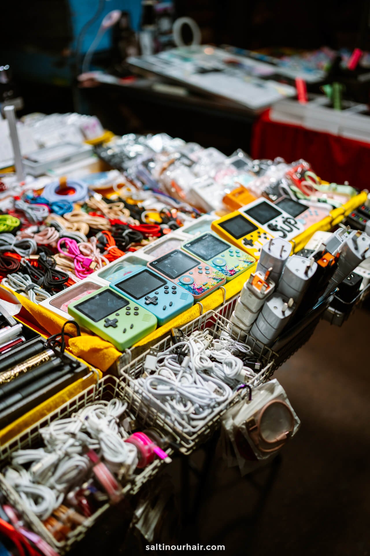 A market stall in Temple Street Market in Hong Kong displays handheld gaming consoles, cords, and electronic accessories neatly organized in wire baskets.