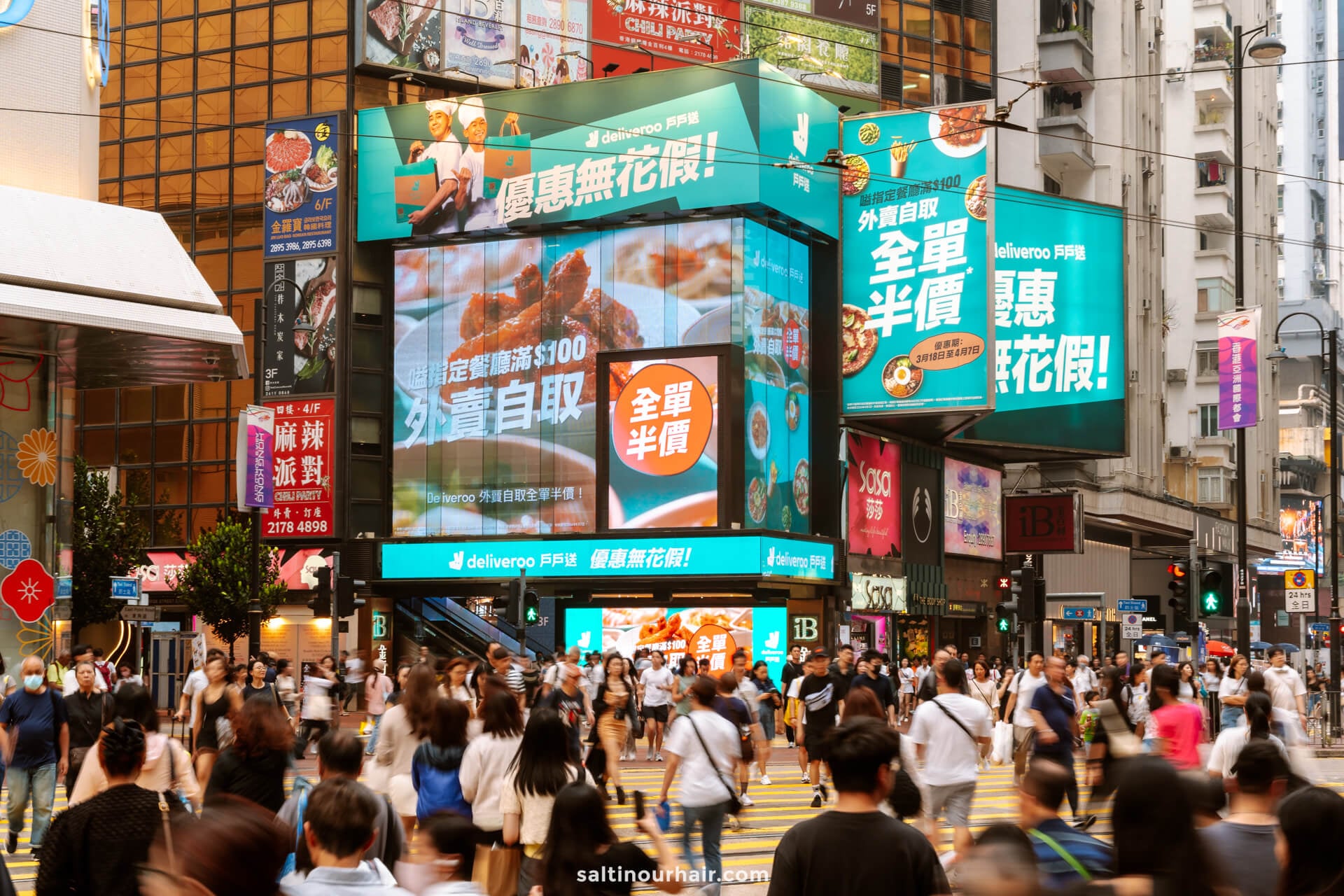 A busy street in Causeway Bay, Hong Kong with large digital and billboard advertisements, crowded with people crossing at a crosswalk