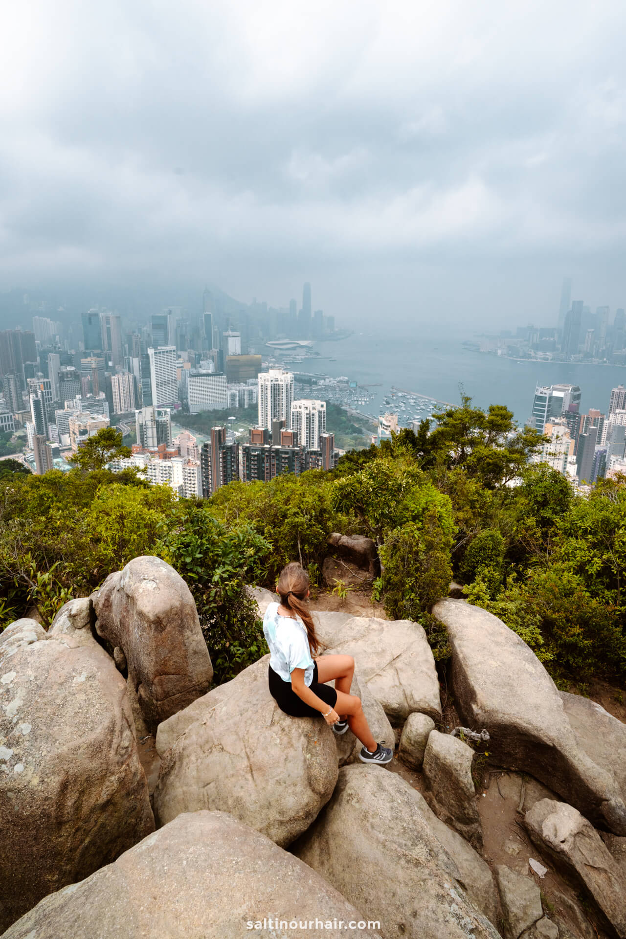 A person sits on large rocks atop a hill, overlooking a city skyline with tall buildings and a body of water under cloudy skies&mdash;a perfect moment to include in your Hong Kong itinerary.