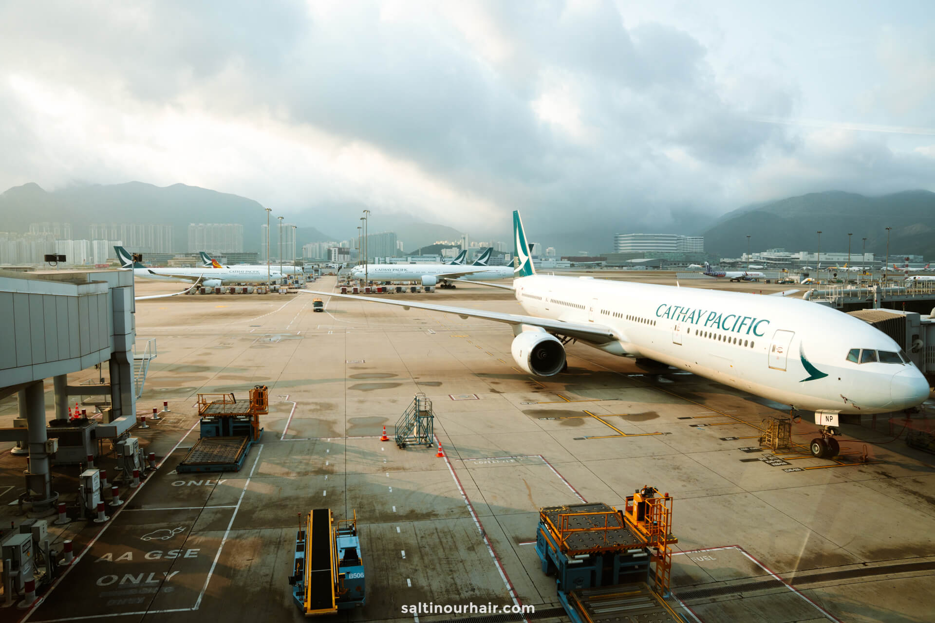 A Cathay Pacific airplane is parked at an airport gate with ground equipment nearby and several other planes in the background