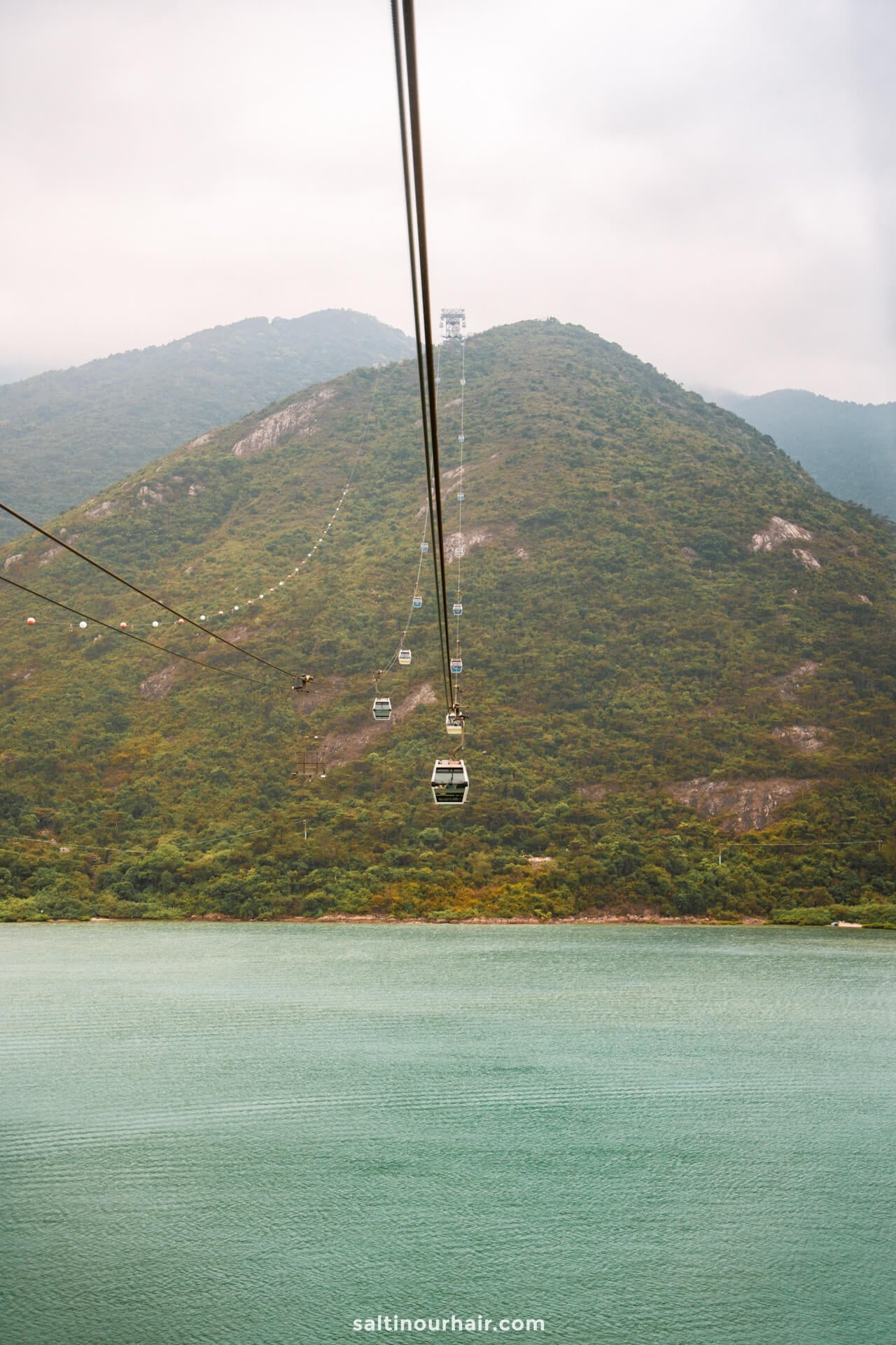 Ngong Ping 360 Cable cars travel above a turquoise body of water toward green, tree-covered hills under a cloudy sky&mdash;a must-see stop on any Hong Kong itinerary.