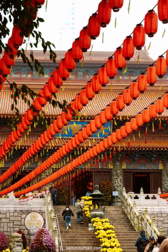Rows of red lanterns are strung above the steps leading to a traditional temple entrance, with people walking and yellow flowers arranged along the path