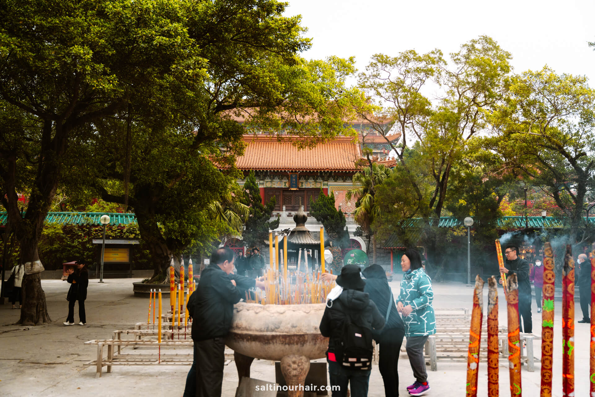 People gather around a large incense burner with tall incense sticks at a temple courtyard, surrounded by trees and traditional architecture in Po Lin Monastery, Hong Kong