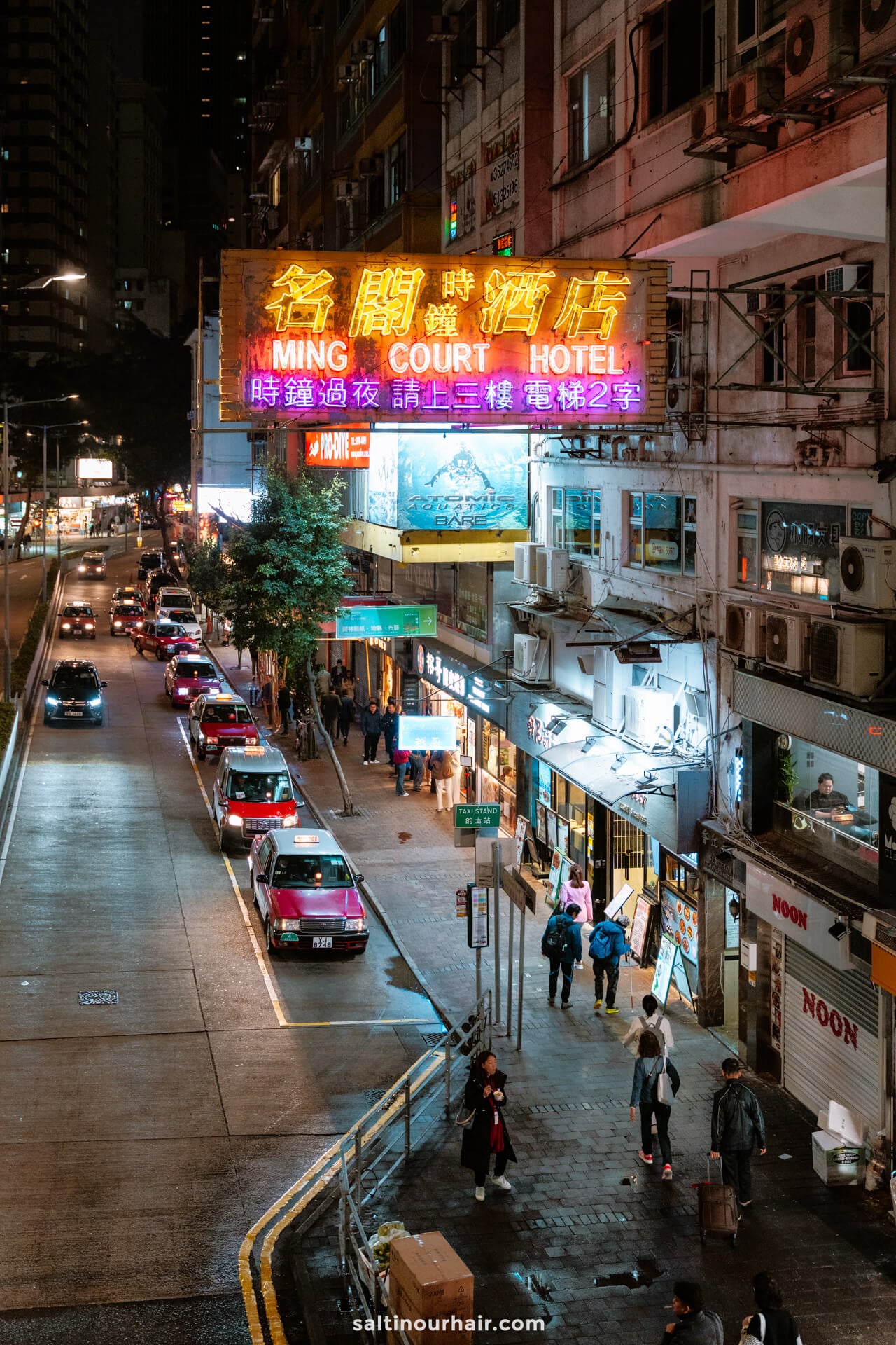 A nighttime city street scene with neon signs, including one for Ming Court Hotel, cars parked along the road, and people walking on the sidewalk.