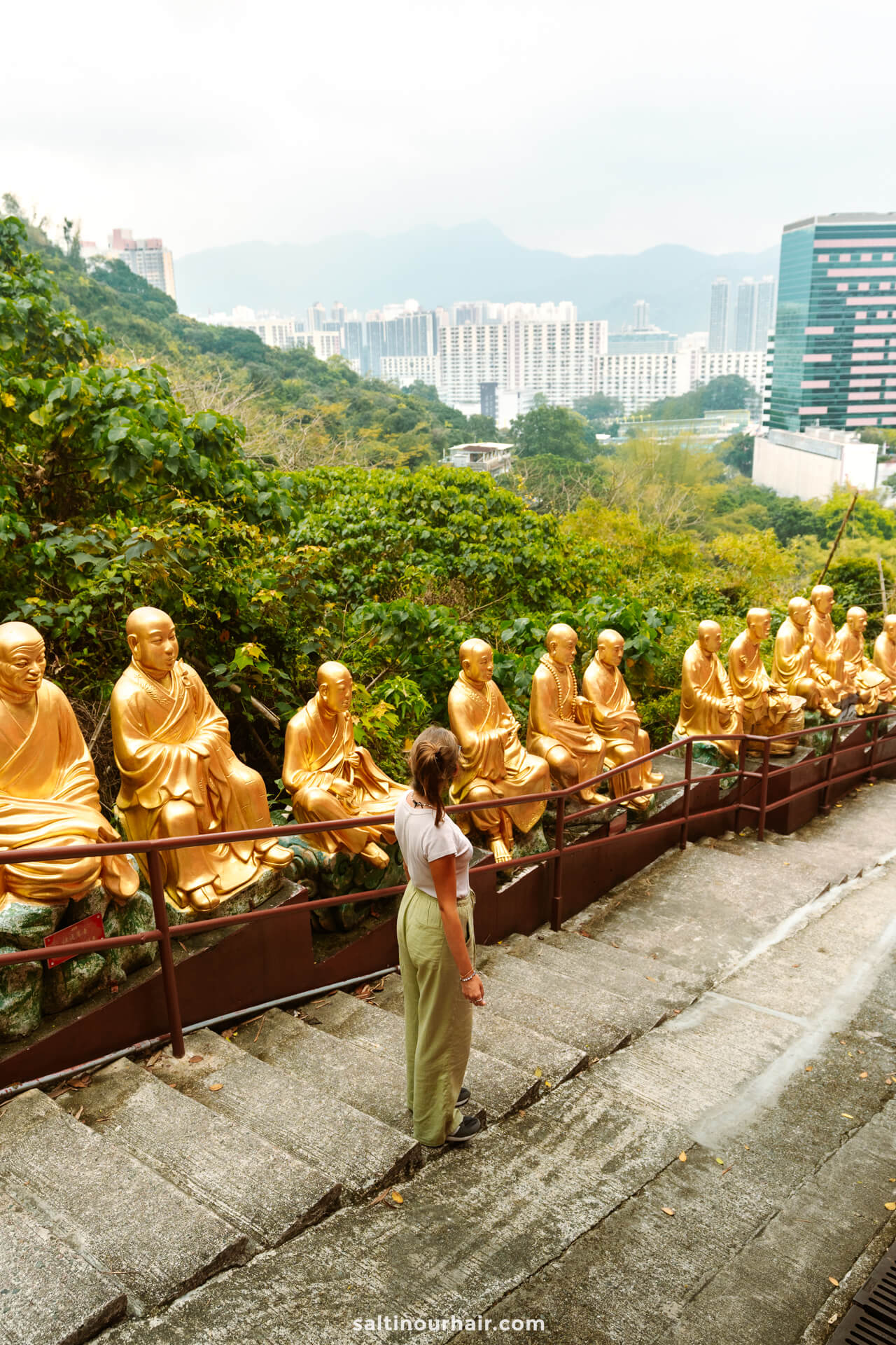 A person stands on stone steps lined with golden monk statues, a unique highlight for any Hong Kong itinerary, with greenery and city buildings visible in the background.
