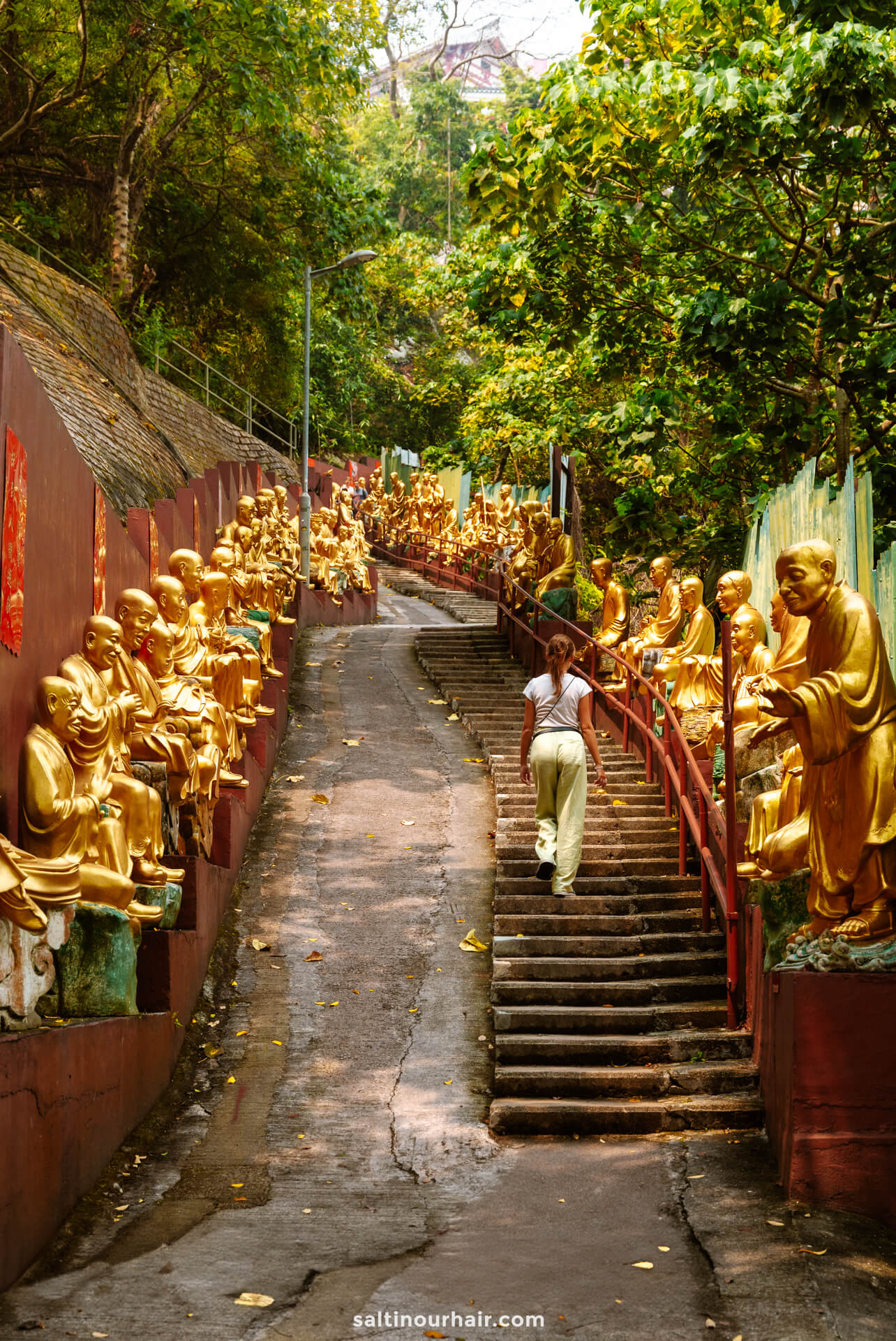 A person walks up a sloped pathway lined with golden Buddha statues on both sides, surrounded by dense green foliage&mdash;a must-see stop on any Hong Kong itinerary.