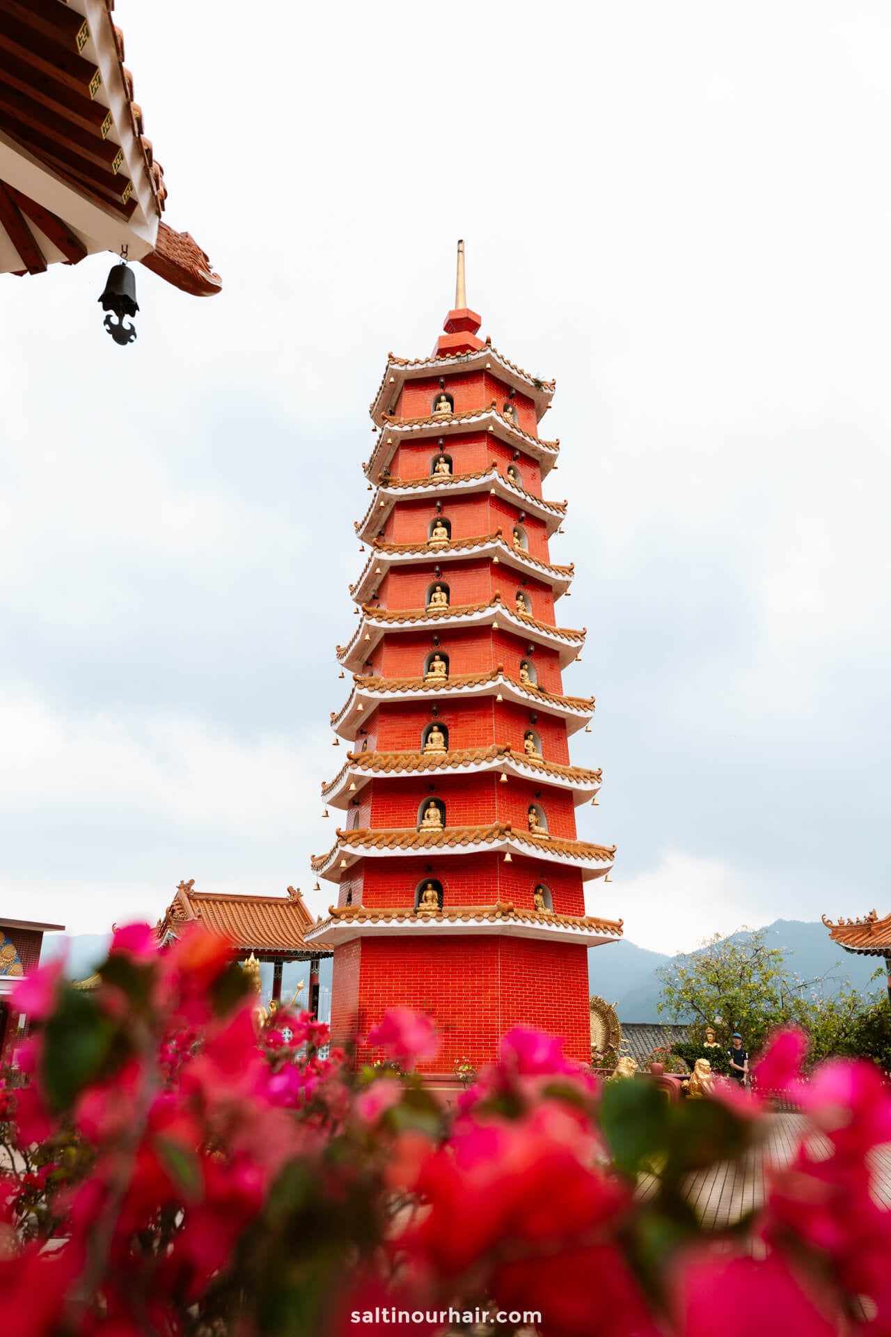 A tall red pagoda with multiple tiers and small Buddha statues, surrounded by pink flowers and set against a cloudy sky at the 10,000 Buddha Monastery in Hong Kong