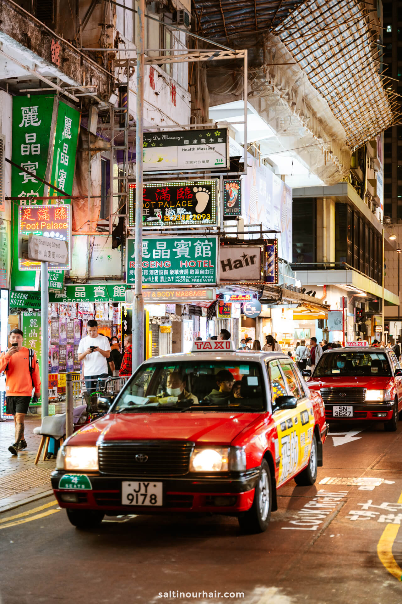 Two red taxis drive down a busy, brightly lit street in Hong Kong lined with neon signs and shops at night