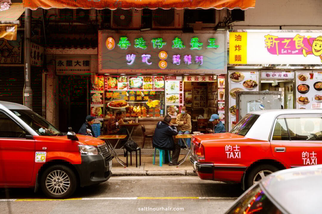 Street scene from a Hong Kong: people sit at tables outside a colorful restaurant with neon signs, flanked by two red taxis parked in front.