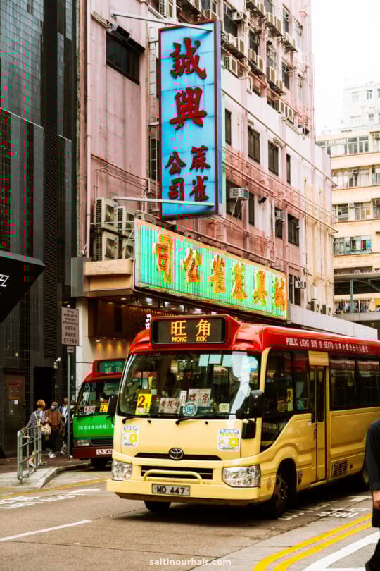 A yellow and red minibus labeled Mong Kok drives through a busy street in Hong Kong, surrounded by tall buildings and neon signs