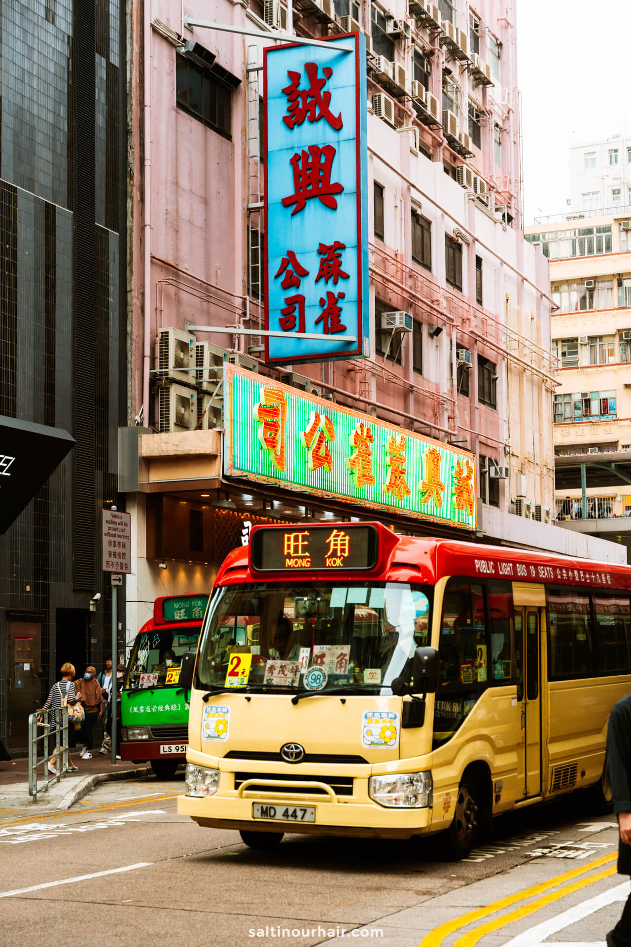 A yellow and red minibus labeled Mong Kok drives through a busy street in Hong Kong, surrounded by tall buildings and neon signs