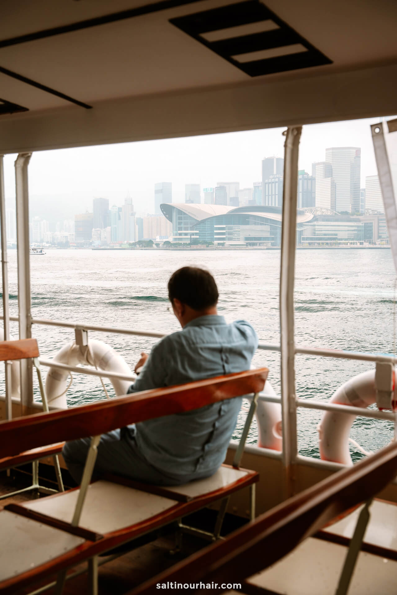 A man sits alone on Star ferry, looking out at the water and city skyline on a cloudy day, perhaps planning his next stop on his Hong Kong itinerary.