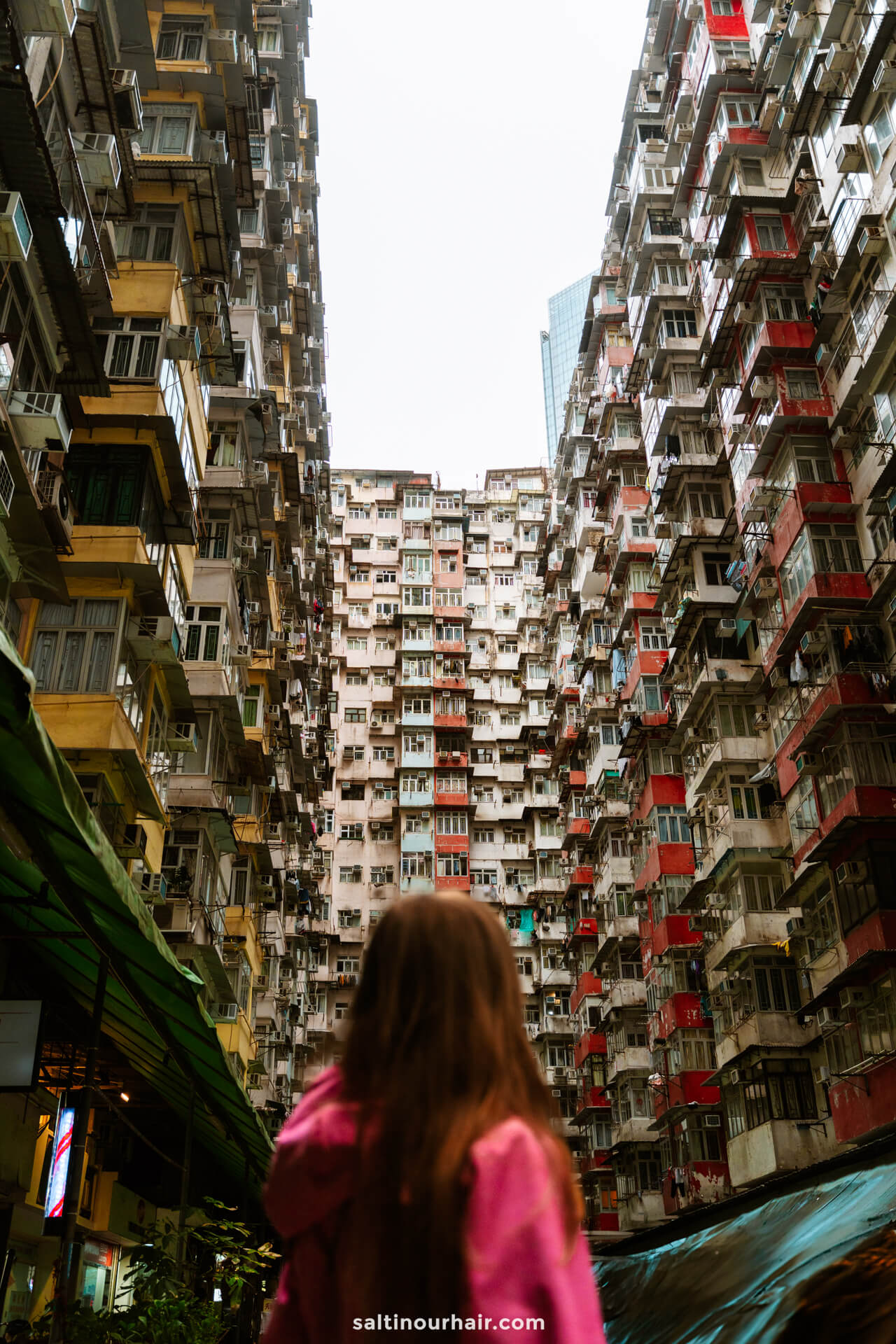A person in a pink hoodie stands facing tall, densely packed apartment buildings with numerous balconies and windows in Hong Kong