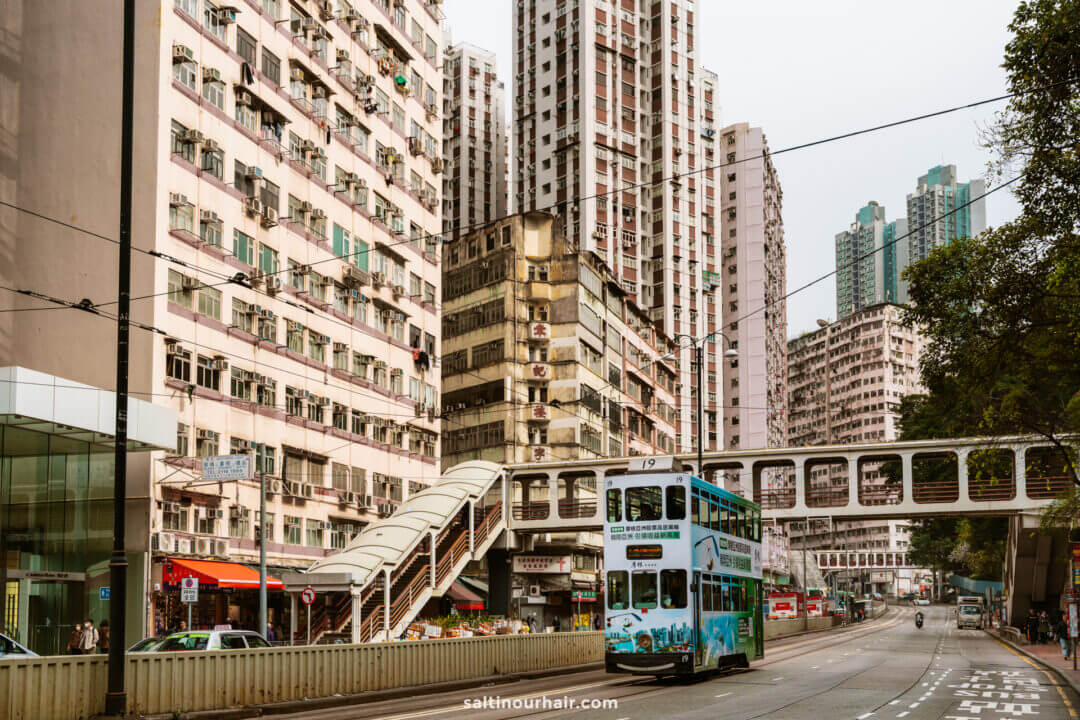 A double-decker tram, a classic feature on any Hong Kong itinerary, passes through a city street lined with tall residential buildings and a pedestrian footbridge.