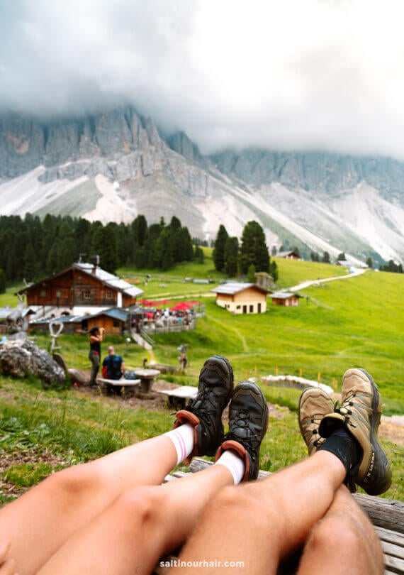Two people with their legs outstretched, wearing hiking shoes, sit on grass overlooking alpine cabins and green hills during the Geisler Alm hike in the Dolomites, with stunning mountains and drifting clouds in the background.