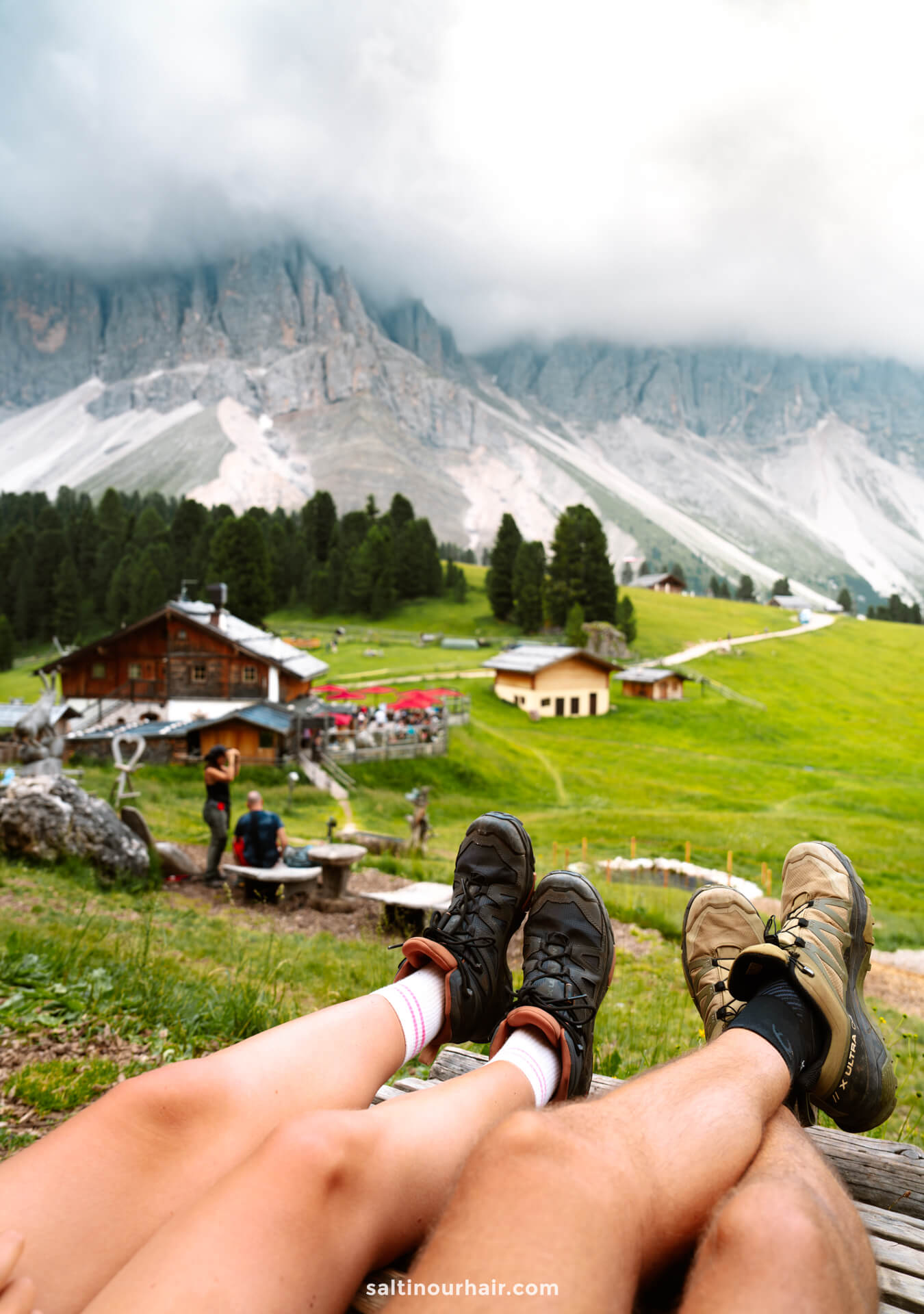 Two people relax with their legs outstretched, overlooking a grassy landscape with the Geisler Alm alpine hut, trees, and cloud-capped mountains—capturing one of the peaceful things to do in the Dolomites.