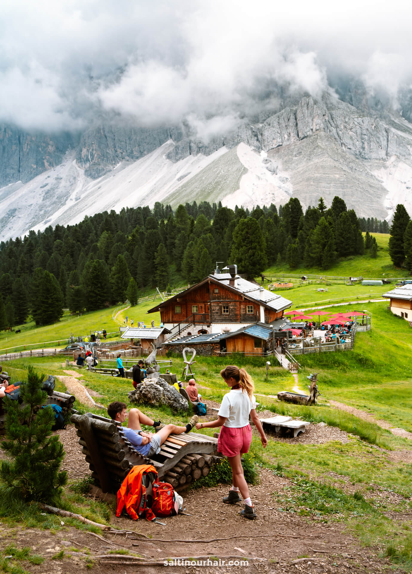 People relax on benches and walk near a rustic wooden cabin called Geisler Alm in a green alpine meadow, surrounded by pine trees and cloudy mountains—one of the many peaceful things to do in the Dolomites.