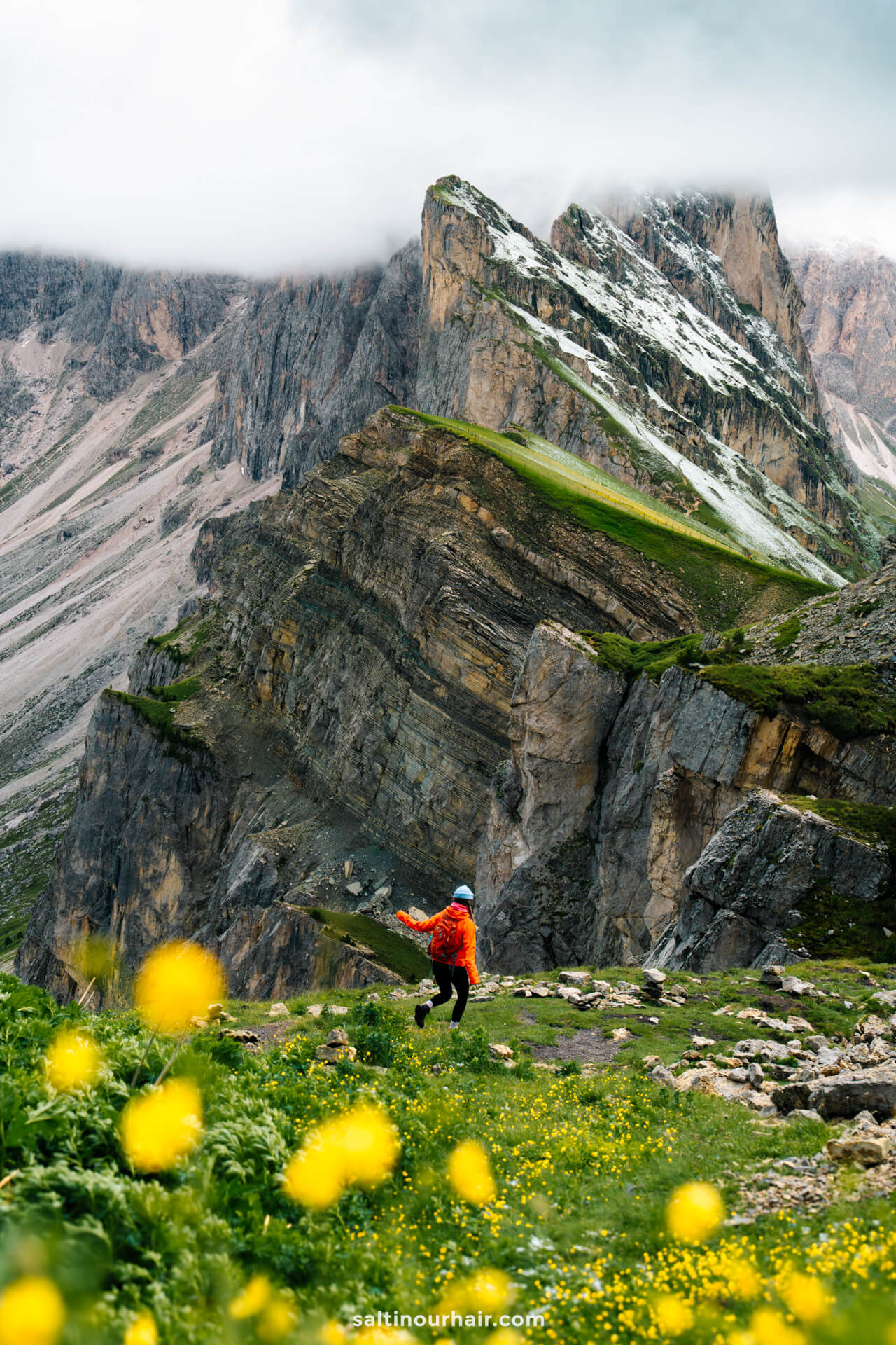 A woman in an orange jacket stands on a rocky mountain path of Seceda surrounded by yellow wildflowers, dramatic cliffs, and clouds—a classic scene among the things to do in the Dolomites.