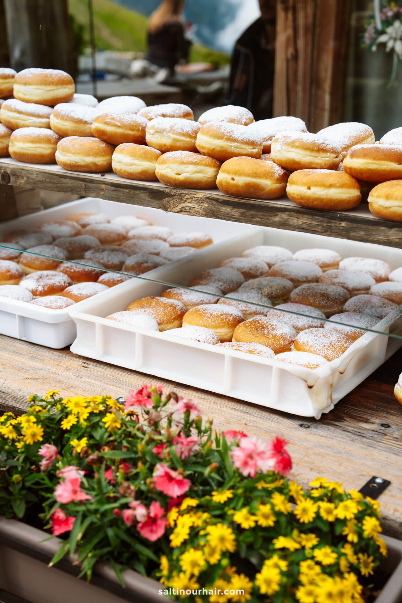 Rows of powdered donuts are displayed on trays and wooden shelves beside colorful flowers at an outdoor market stall, a delightful stop among the many things to do in the Dolomites.