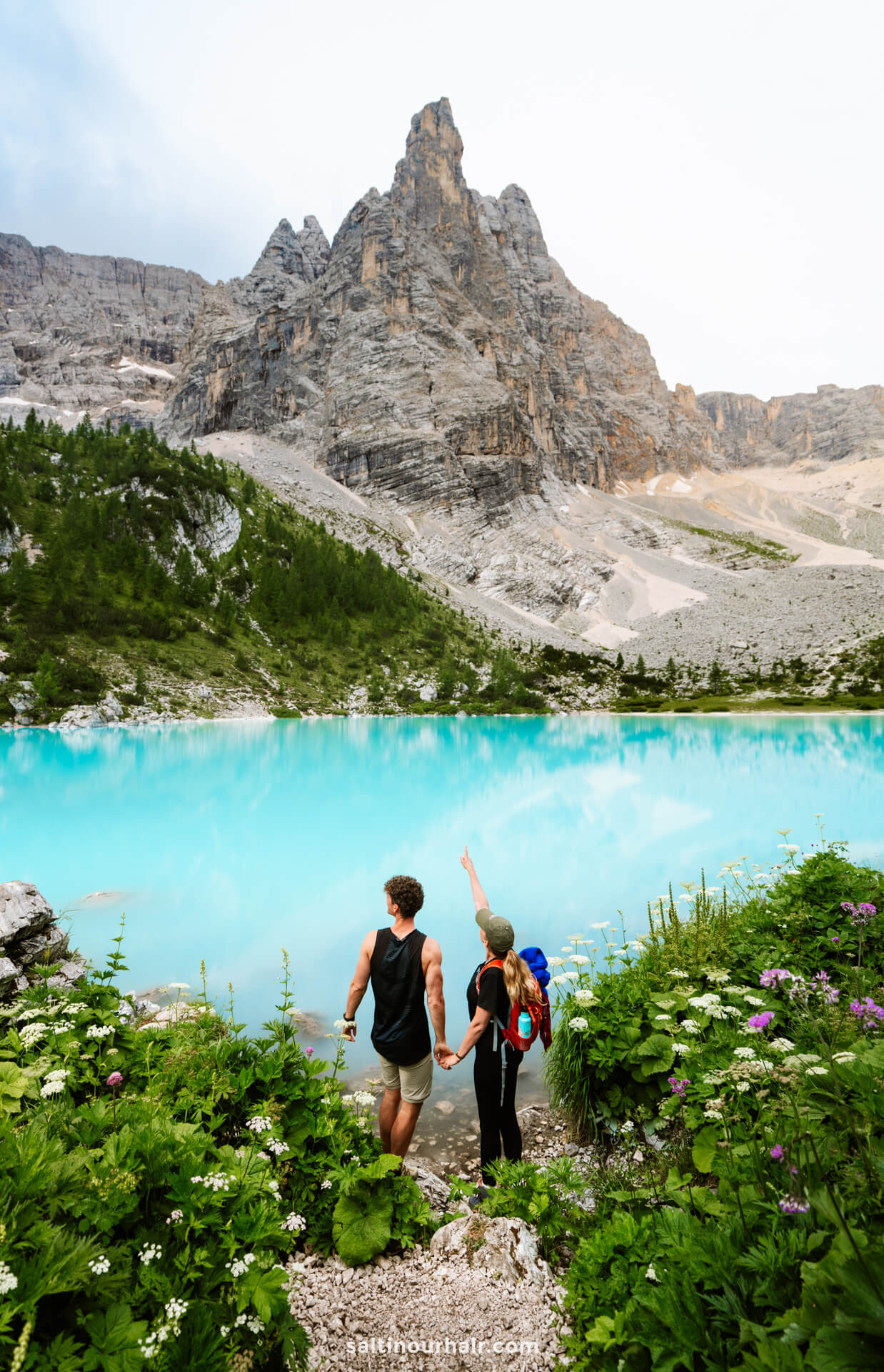 Two people holding hands stand by a turquoise lake called Lago di Sorapis with lush greenery and wildflowers, facing a jagged mountain peak under a cloudy sky; one points toward the summit, dreaming of new things to do in the Dolomites.