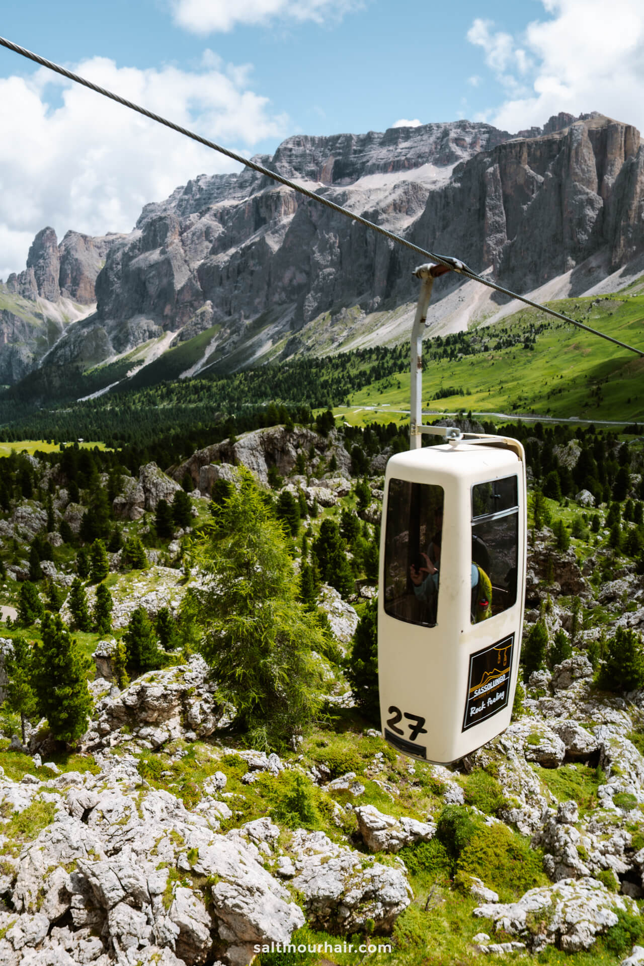 A white coffin cable car glides above rocky, green alpine terrain with tall mountains in the background under a partly cloudy sky—one of the top things to do in the Dolomites.