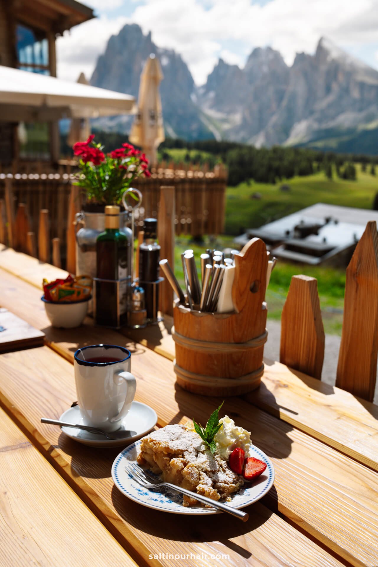 A cup of coffee and a slice of cake with fruit on a wooden table outdoors, overlooking scenic mountain landscapes of alpe di siusi—one of the delightful things to do in the Dolomites italy