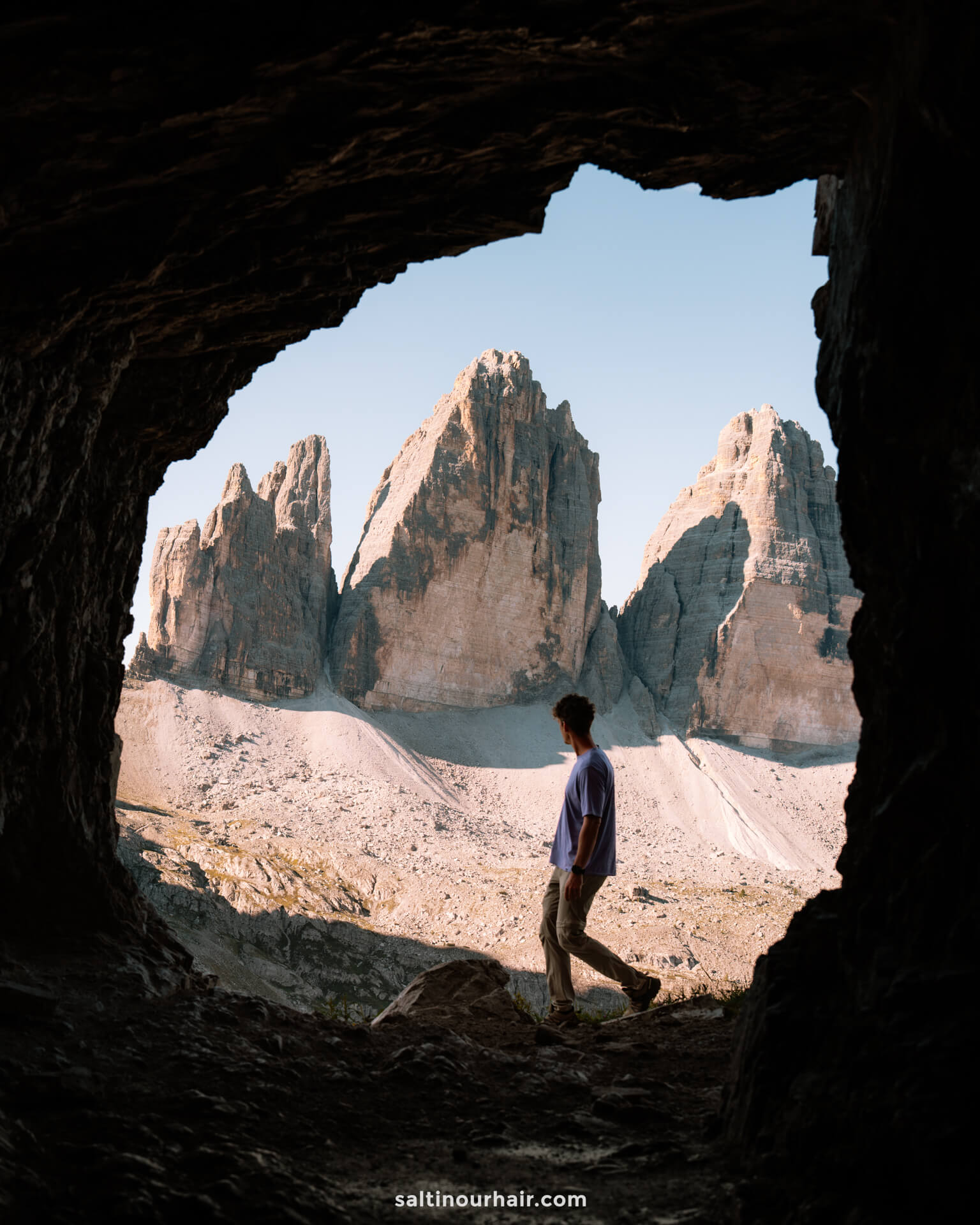 A person stands at the entrance of a rocky cave, looking at three large mountain peaks called Drei zinnen under a clear sky—one of the many unforgettable things to do in the Dolomites.