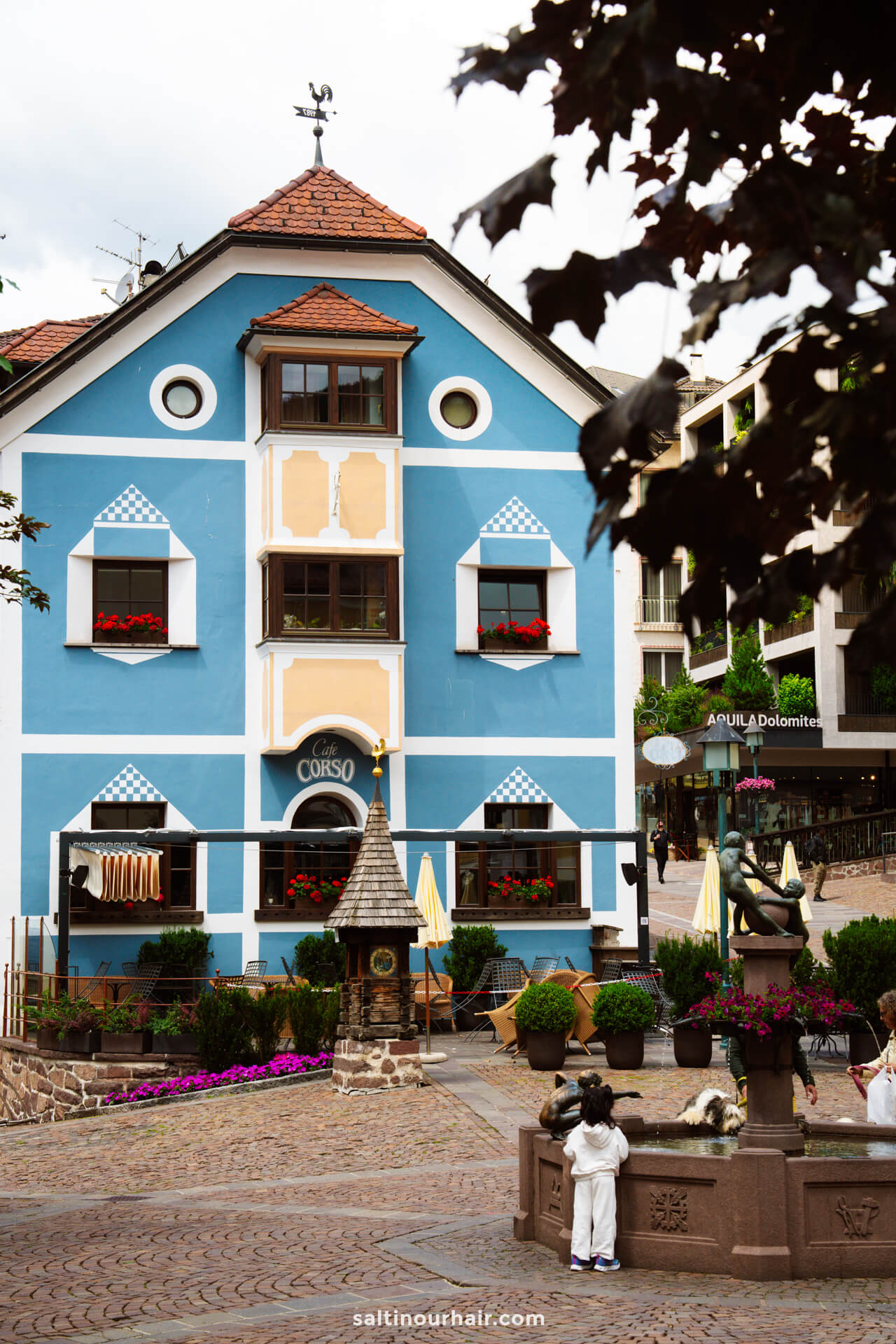 A blue and white building with flower boxes and striped awnings stands in a cobblestone plaza; a child looks at a bronze statue in the foreground—a charming scene among the many things to do in the Dolomites italy