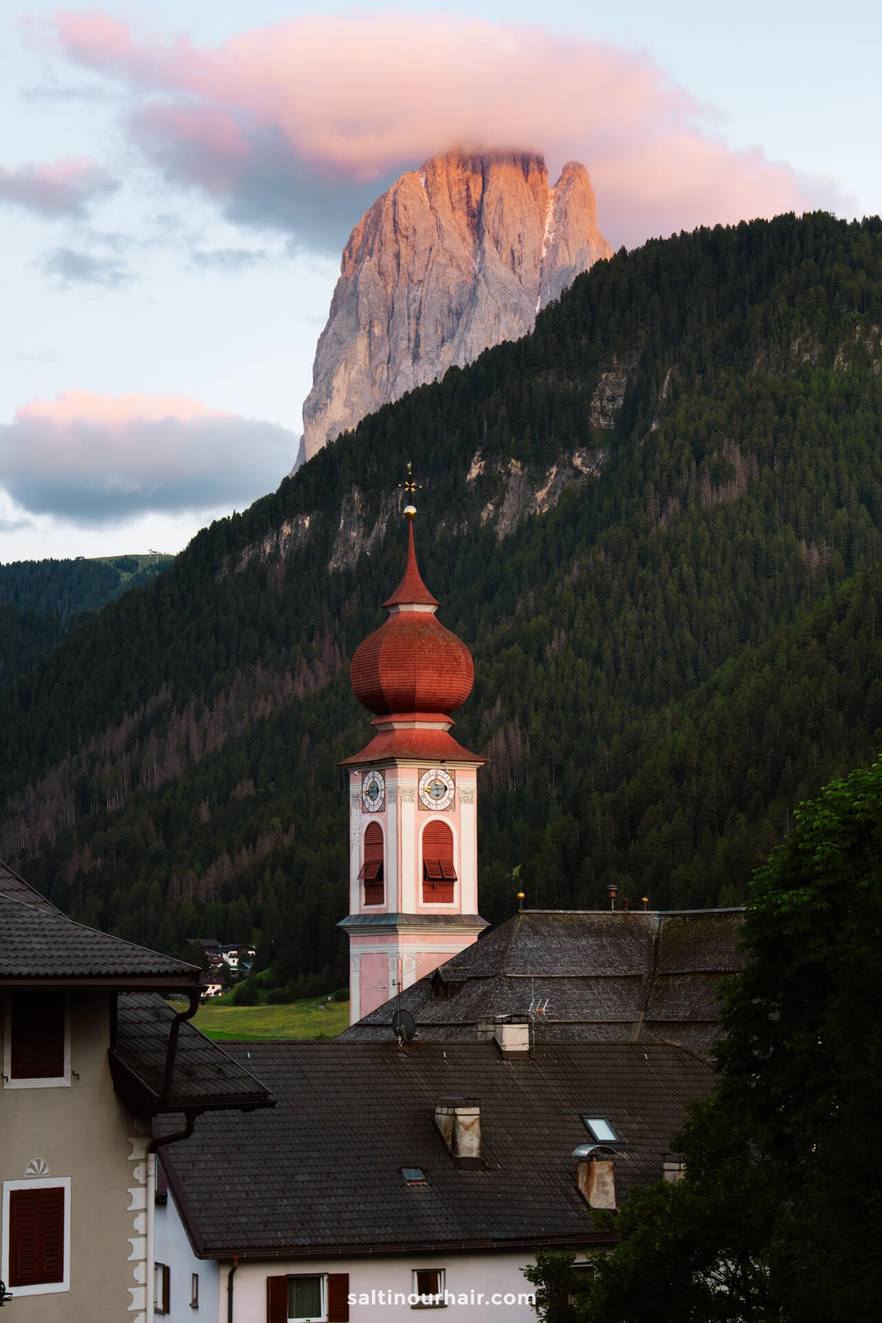 A red-domed church tower in Ortisei stands before a forested hill, with a rocky mountain peak and pink clouds in the background—capturing the breathtaking scenery among the top things to do in the Dolomites.