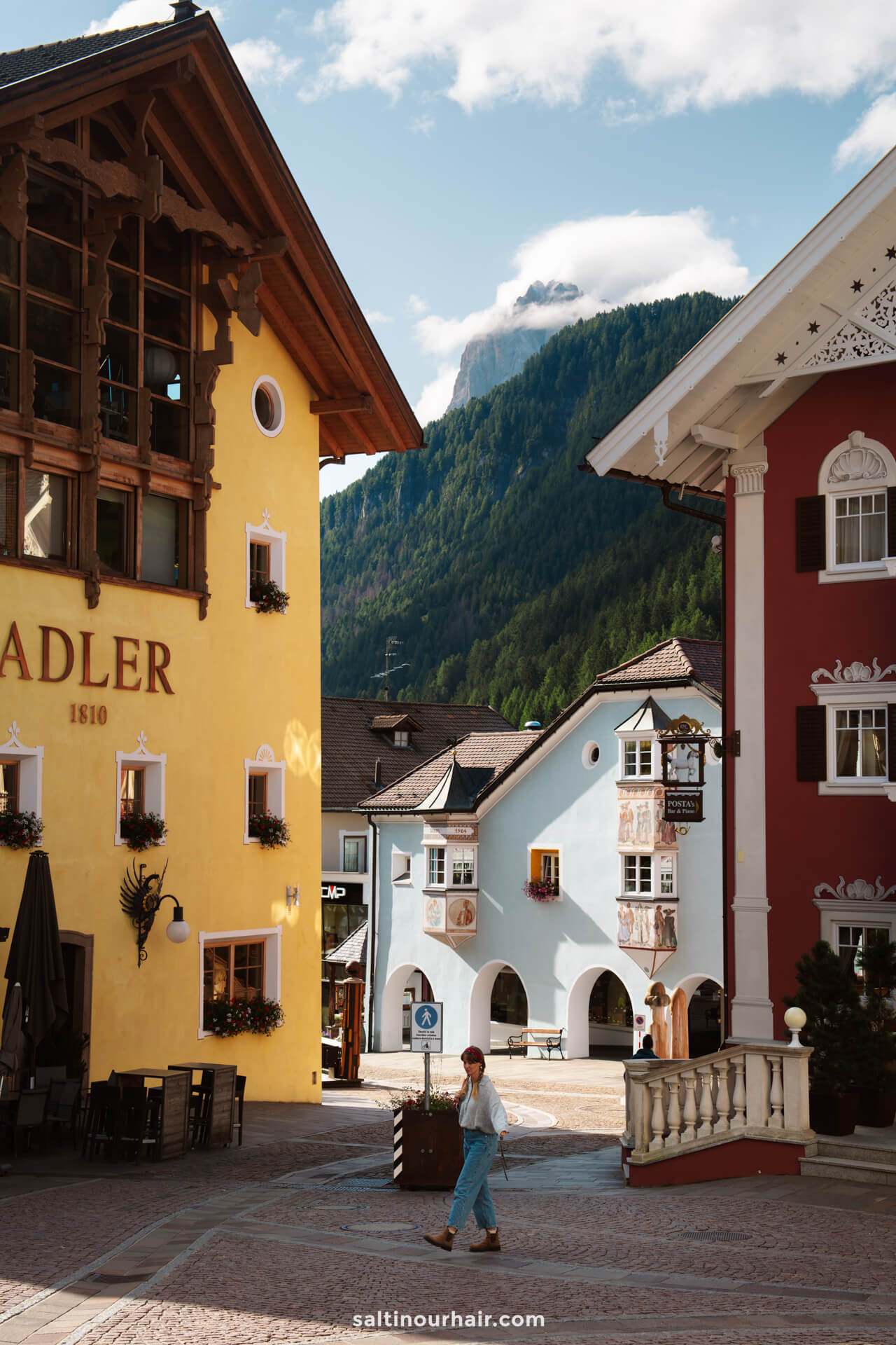 A woman walks through the quaint Ortisei town square with colorful buildings, mountains in the background, and outdoor seating visible—one of many charming things to do in the Dolomites italy