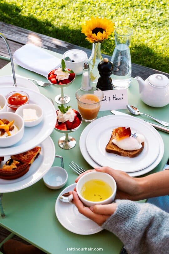 A table set outdoors, perfect for relaxing after exploring things to do in the Dolomites, features tea, a poached egg on toast, desserts in glass cups, condiments, a glass water bottle, and a sunflower in a vase. A person holds a cup of tea.