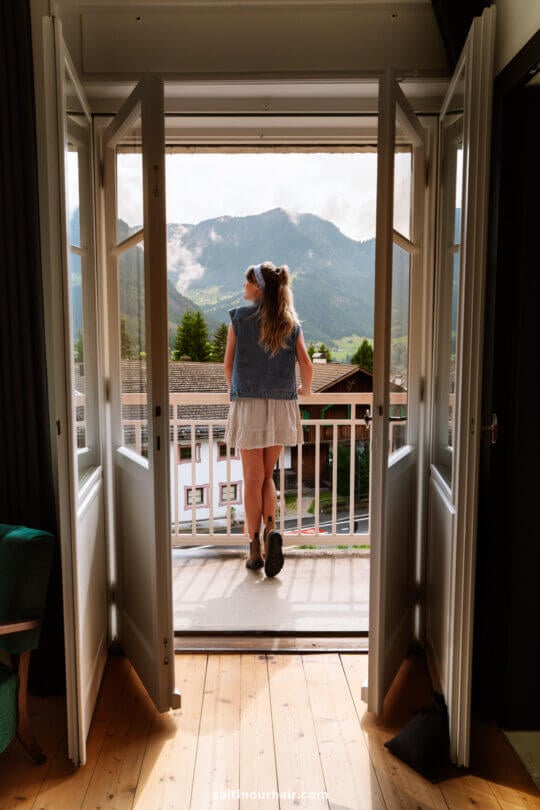A woman in a white dress and denim vest stands on a balcony at hotel Casa al Sole in Ortisei, looking out at a mountain landscape through open double doors, dreaming of things to do in the Dolomites.