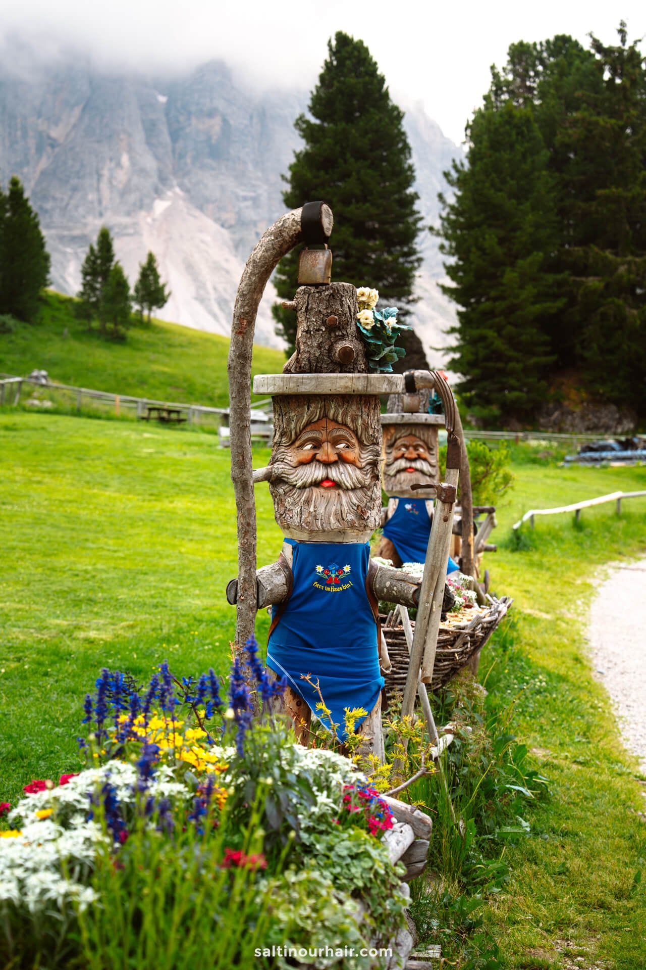 Wooden gnome statues wearing blue aprons stand along a path in a grassy, mountainous landscape with flowers&mdash;one of the charming things to see at the Geisler alm hut in dolomites italy