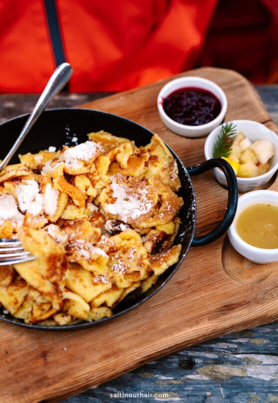 A skillet of Kaiserschmarrn topped with powdered sugar, served on a wooden board with sides of fruit compote, diced fruit, and sauce—an authentic treat among the best things to do in the Dolomites.