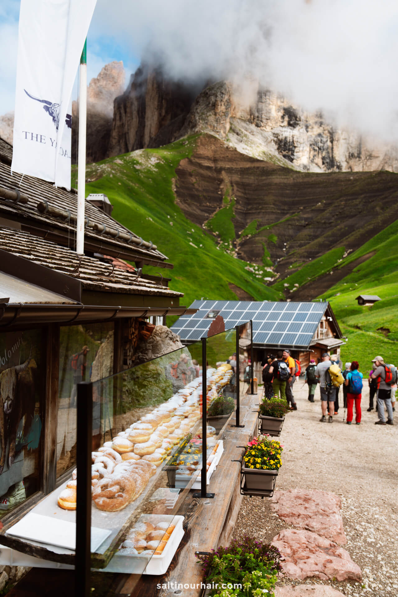 Glass display of donuts outside a mountain cafe; hikers stand nearby, enjoying one of the best things to do in the Dolomites, with green hills and rocky peaks in the background under a cloudy sky.