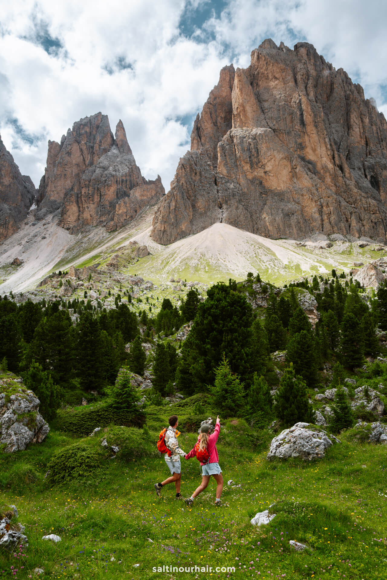 Two hikers with backpacks walk through a grassy, rocky area dotted with evergreen trees, approaching the jagged peaks—a perfect scene for those seeking things to do in the Dolomites under a dramatic, cloudy sky.