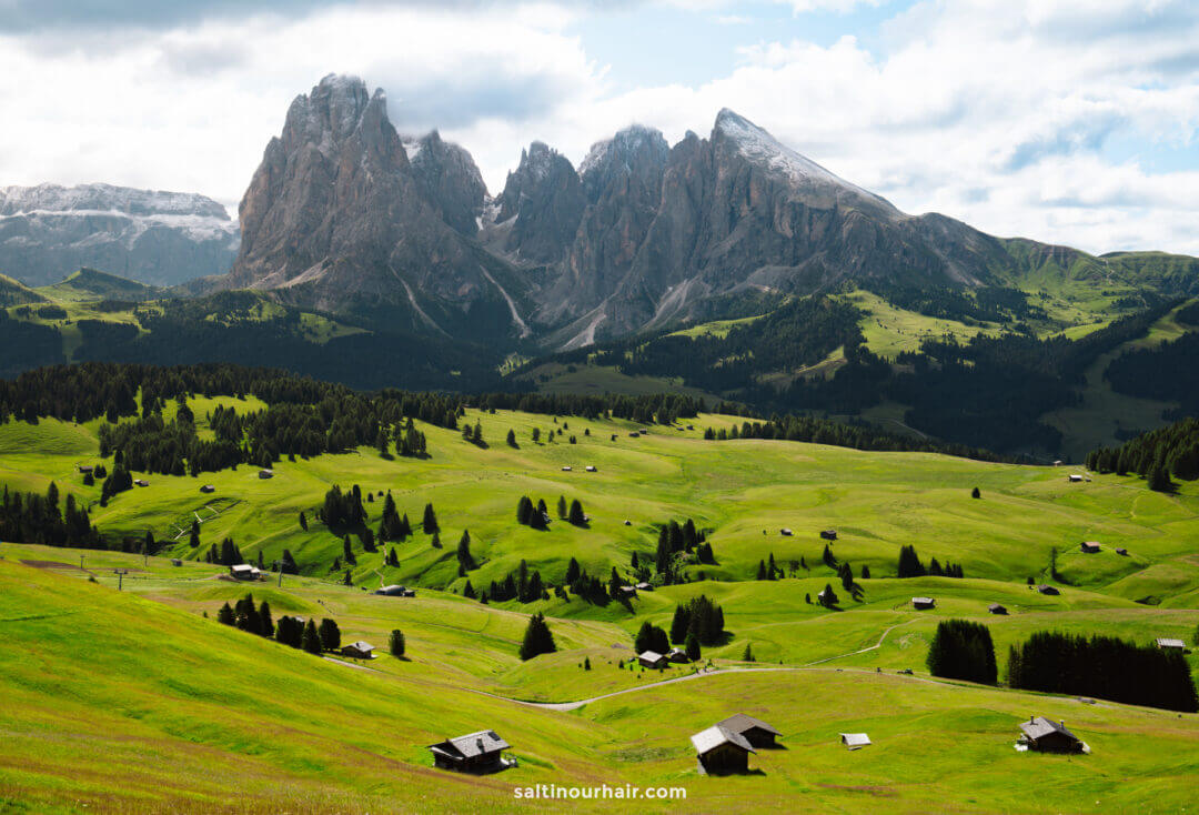 Green alpine meadows called alpe di siusi dotted with wooden huts in the foreground and rugged, rocky mountains rising behind hint at the many things to do in the Dolomites under a partly cloudy sky.