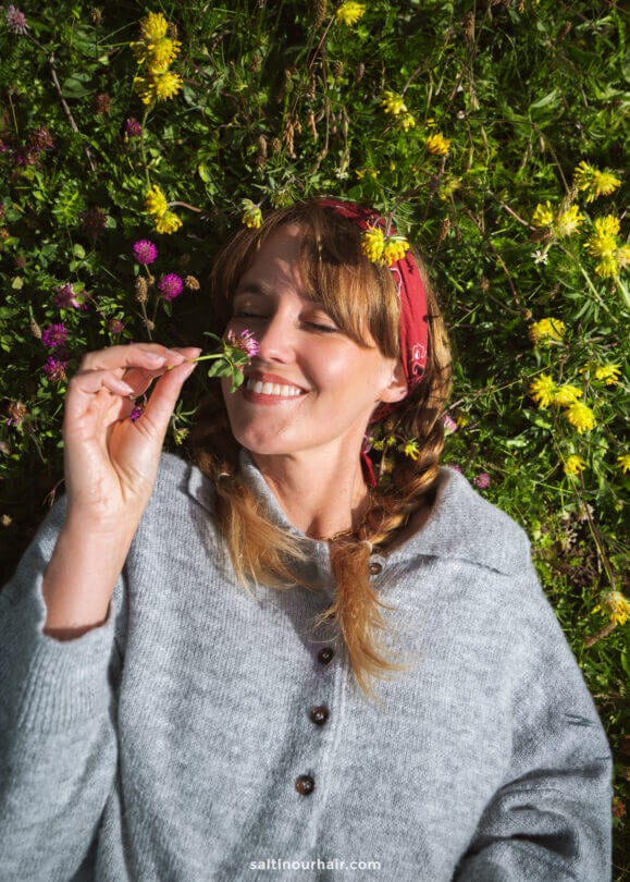 A woman with braided hair and a red bandana lies on grass dotted with yellow and purple flowers, holding a clover bloom close to her face and smiling—capturing the joy of simple things to do in the Dolomites.