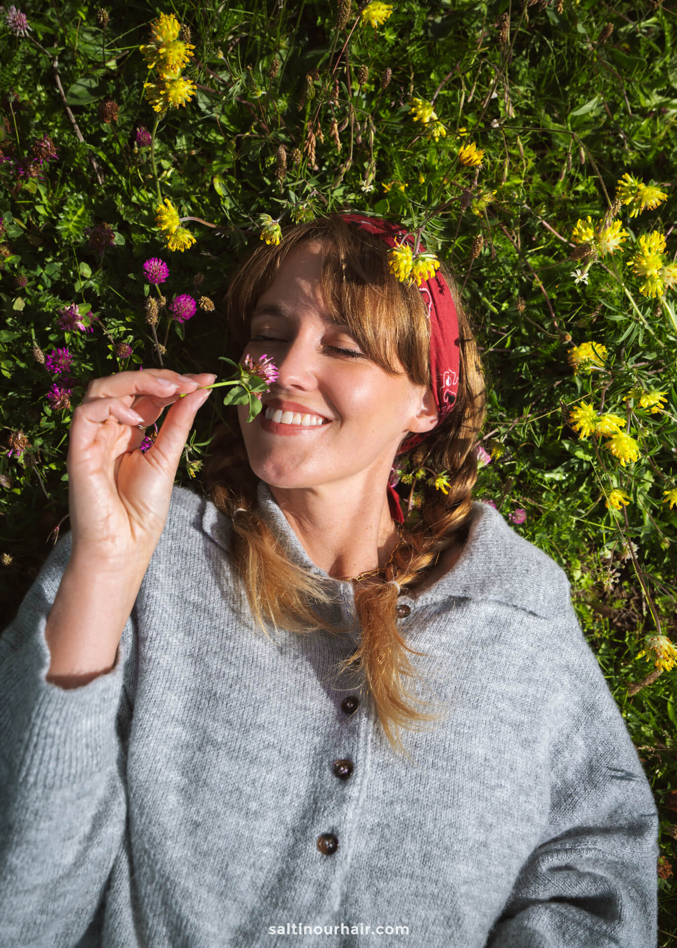 A woman with braided hair and a red bandana lies on grass with wildflowers in Alpe di Siusi, Dolomites, Italy, smiling and holding a small flower near her nose.