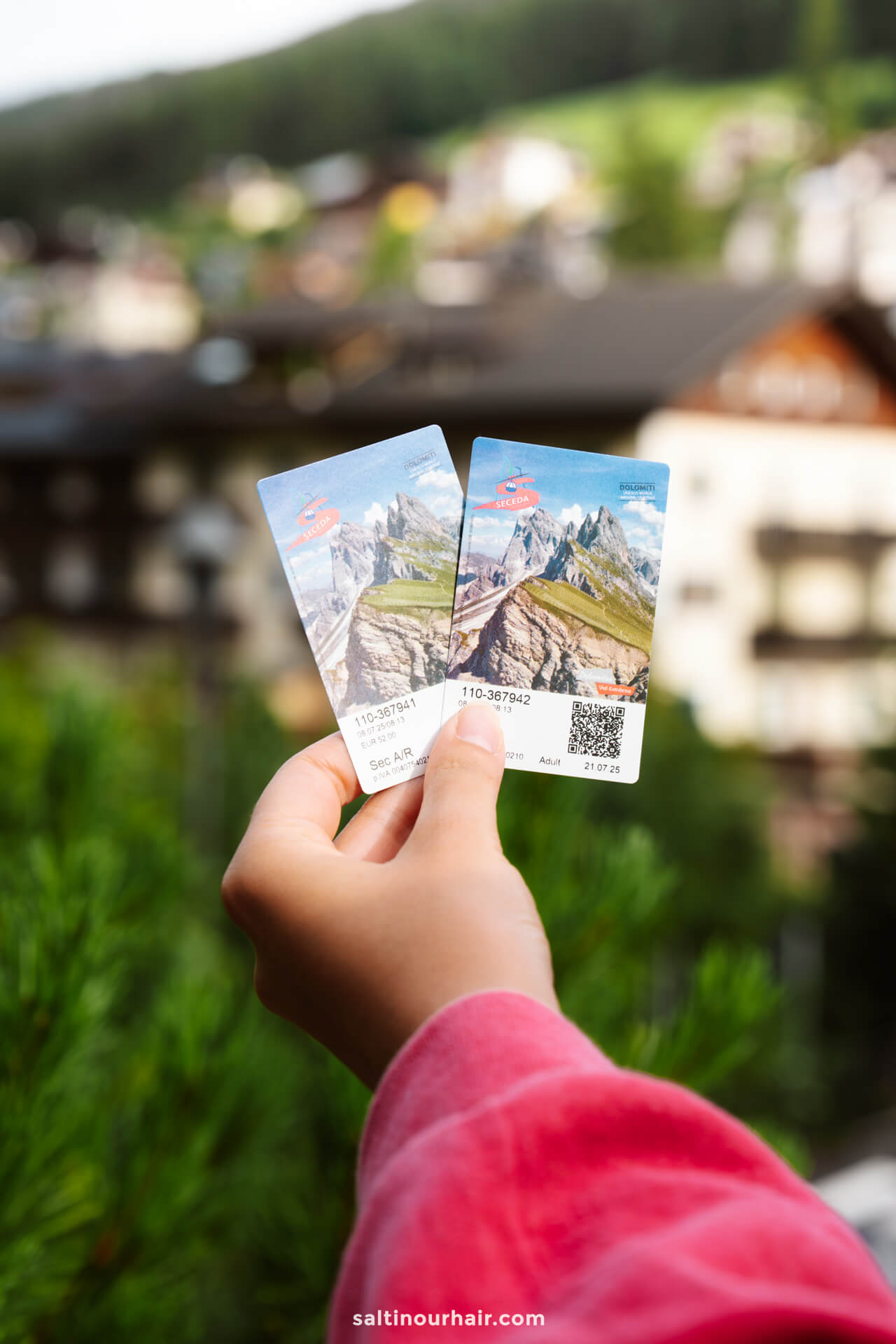 A hand holds two scenic mountain cable car tickets with a blurred background of buildings and trees—one of the must-try things to do in the Dolomites.