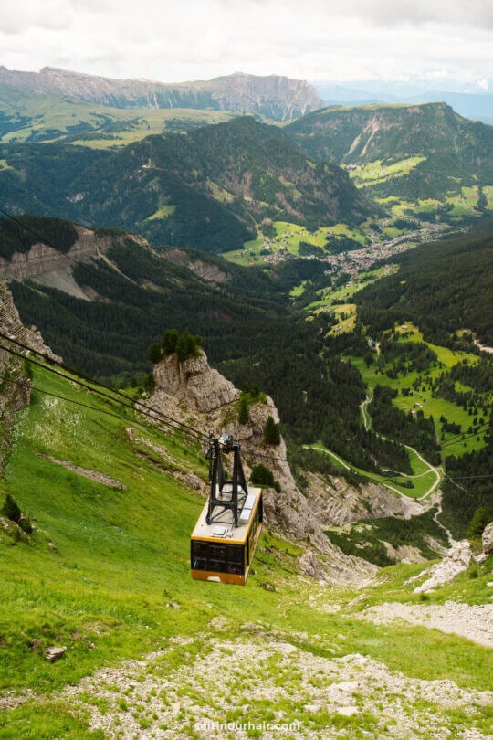 A yellow cable car ascends a steep mountain slope at Seceda, offering breathtaking views of a lush green valley, forests, and distant villages below—one of the unforgettable things to do in the Dolomites.